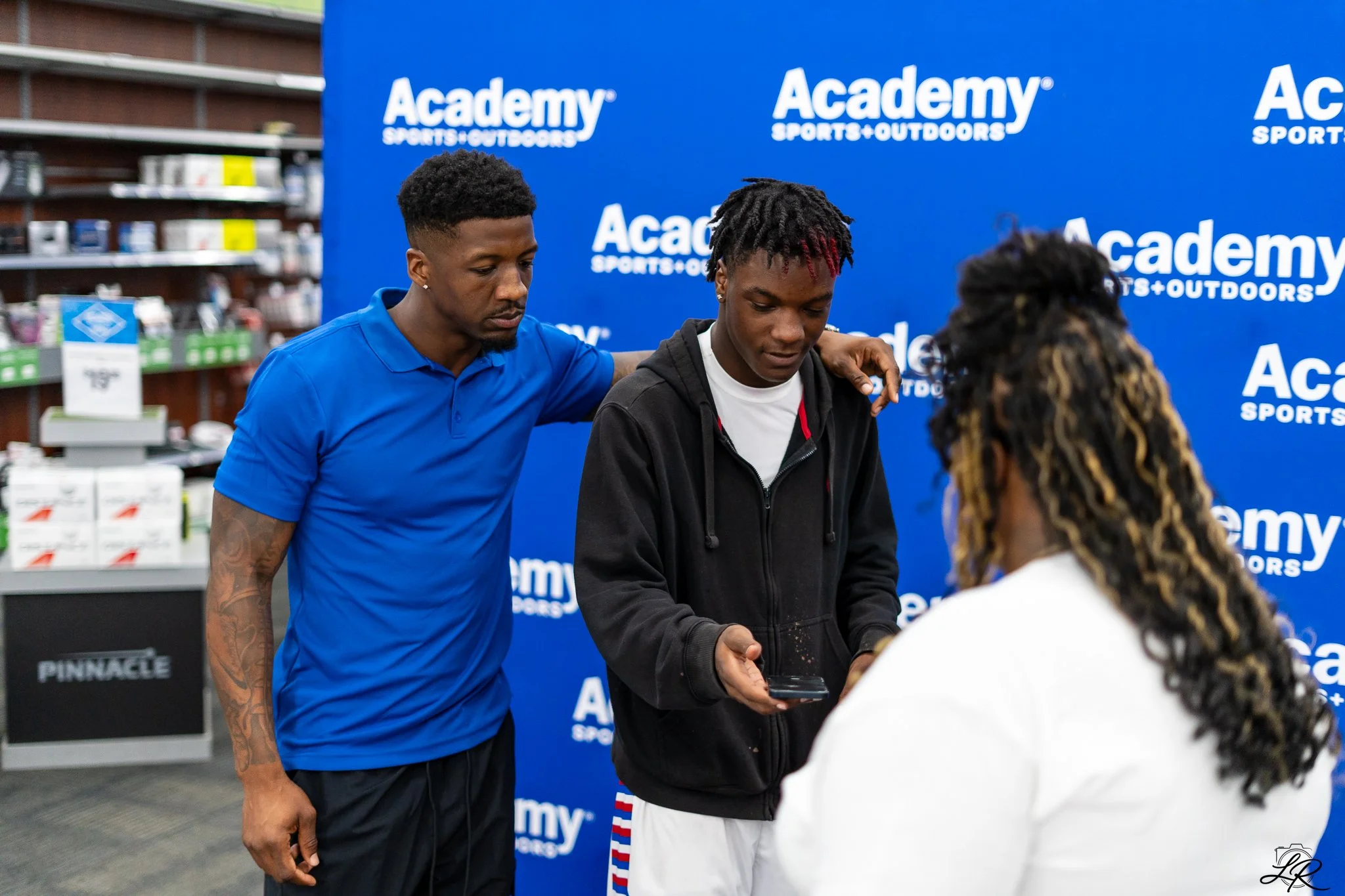 Two young men are shaking hands inside a store with a blue backdrop that reads 'Academy Sports + Outdoors.' One man is wearing a blue polo shirt, and the other is in a black hoodie with red highlights in his hair. There is a third person with long cu
