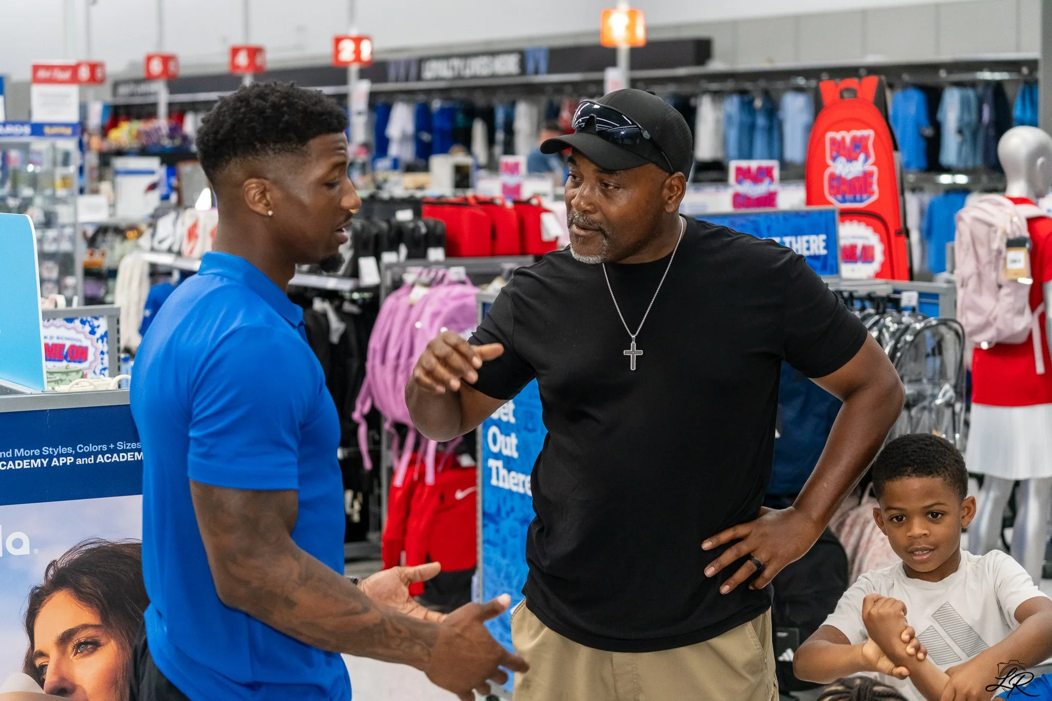 Two men shaking hands in a retail store, with children nearby and shelves filled with backpacks and apparel in the background.