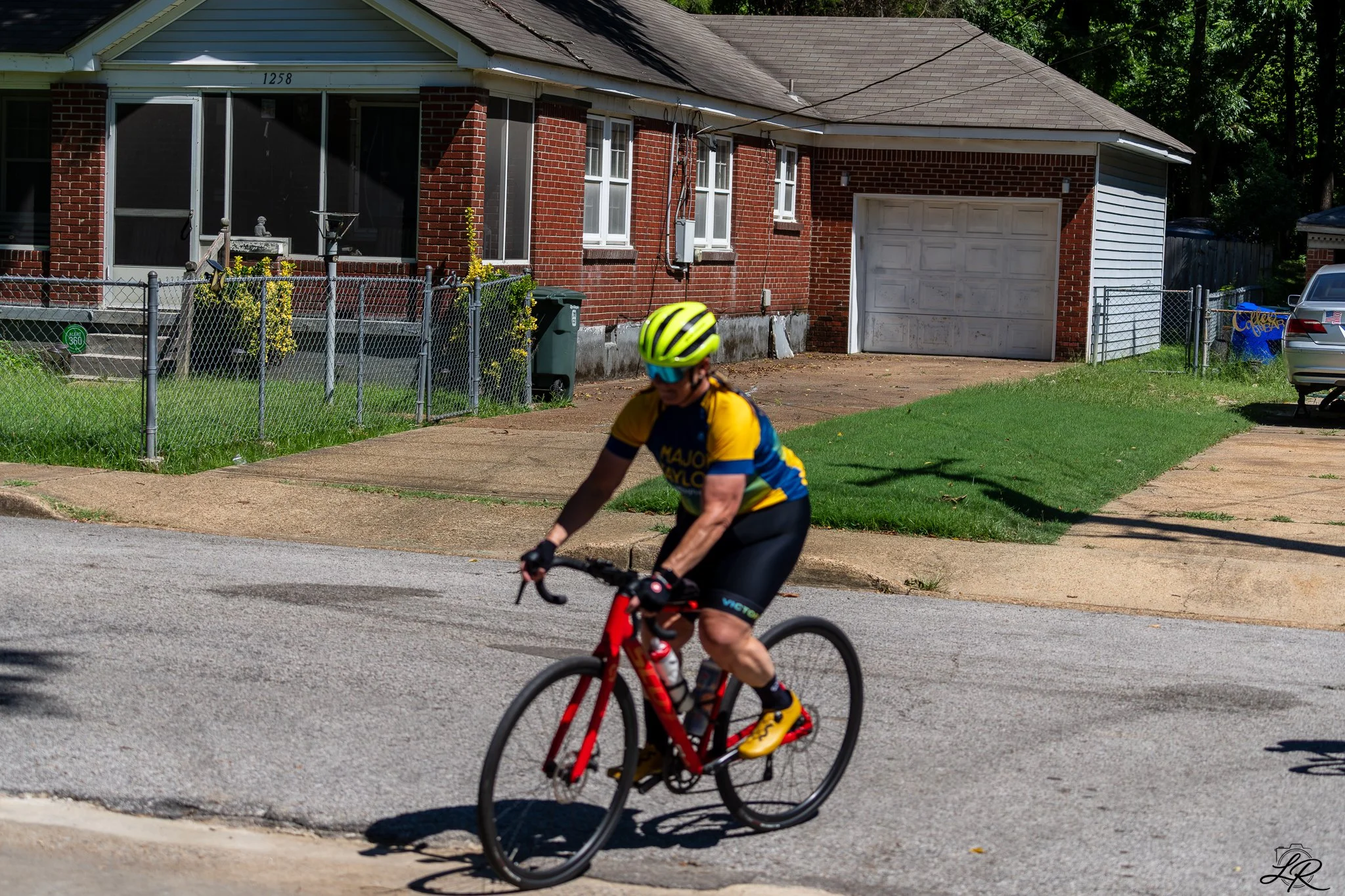 A person wearing a yellow and black cycling uniform and a yellow helmet riding a red bicycle on a suburban street with a brick house and a fenced yard in the background.