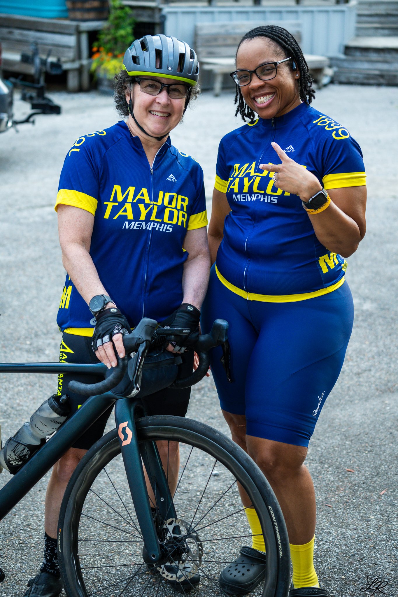 Two women wearing blue and yellow cycling jerseys with the words "Major Taylor Memphis" stand outdoors. One woman is holding a bike and wearing a helmet, and the other is smiling and pointing. They are smiling and appear to be enjoying a sunny day.