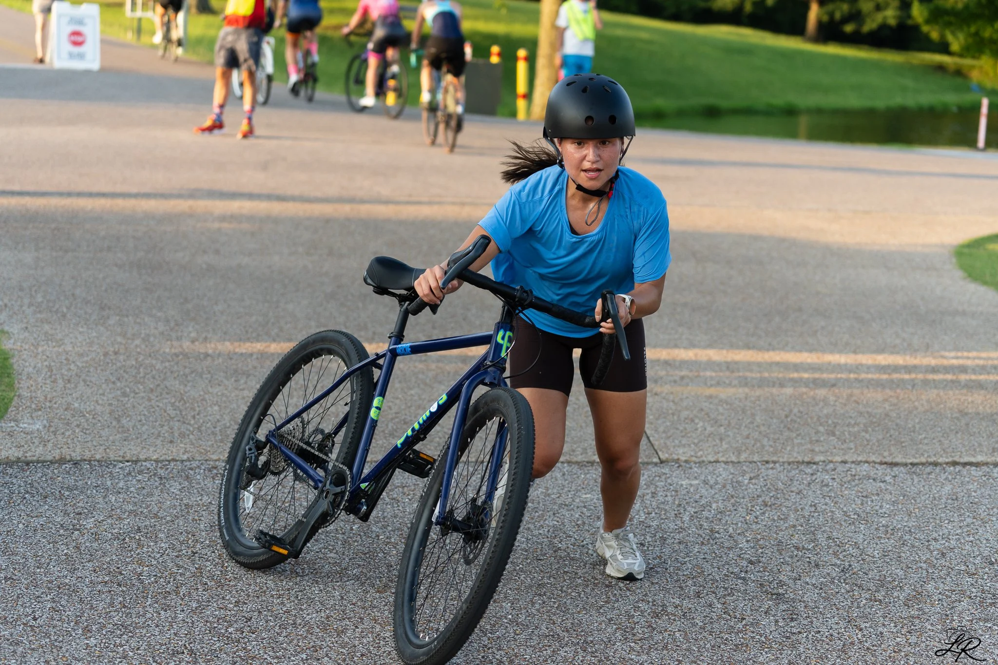 A young woman in a blue athletic shirt, black helmet, and shorts pushing a mountain bike on a paved path, with a park and people in the background, some on bicycles and others walking.