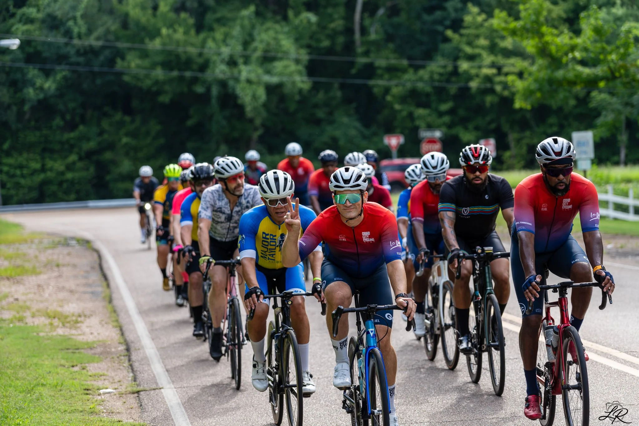 A large group of cyclists riding on a tree-lined road. They are wearing helmets and colorful cycling jerseys.
