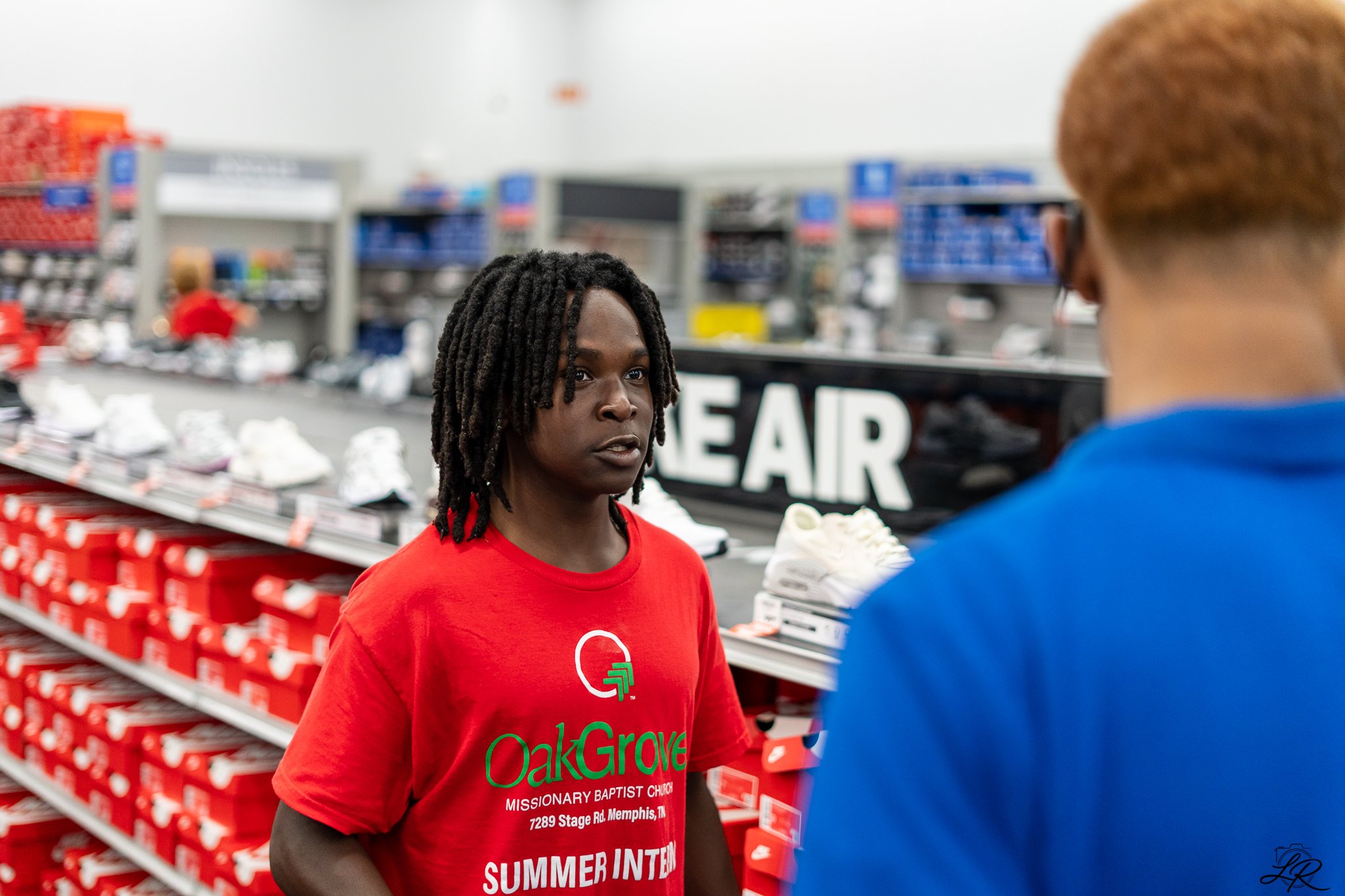 A woman with dreadlocks wearing a red t-shirt with 'OakGrove Missionary Baptist Church' printed on it, standing in a store aisle facing a person with short hair and glasses. The store background includes shelves of shoes and boxes, with a partially v
