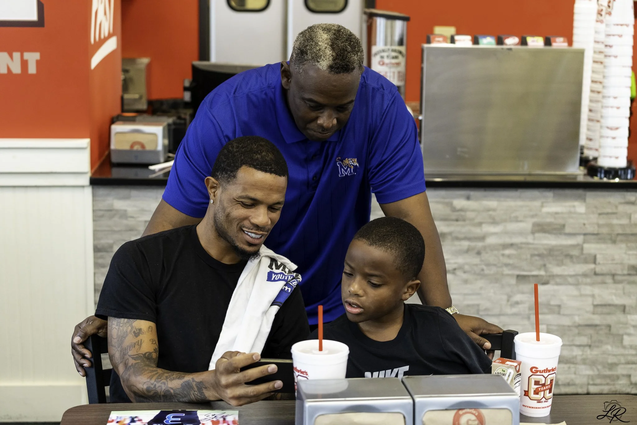 A man with tattoos on his left arm and a woman are sitting at a table with a young boy, all looking at a smartphone that the man is holding. Behind them, a man in a blue shirt with an 'M' logo is leaning over and looking at the phone as well. There a