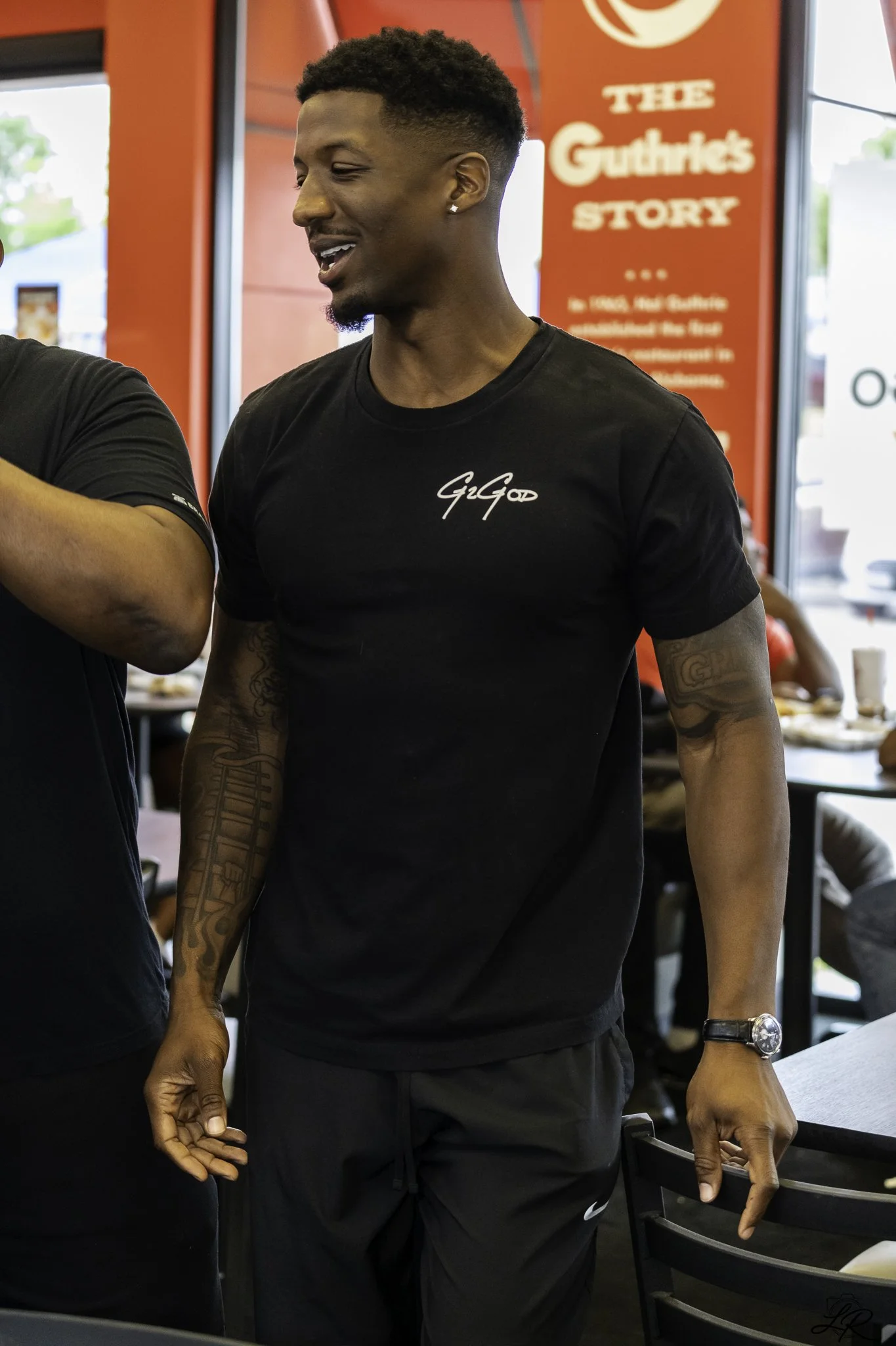 Young man with short curly hair, earrings, and tattooed arms, smiling and holding hands with someone, inside a restaurant or cafe with a red sign in the background reading 'The Gurkhas Story'.