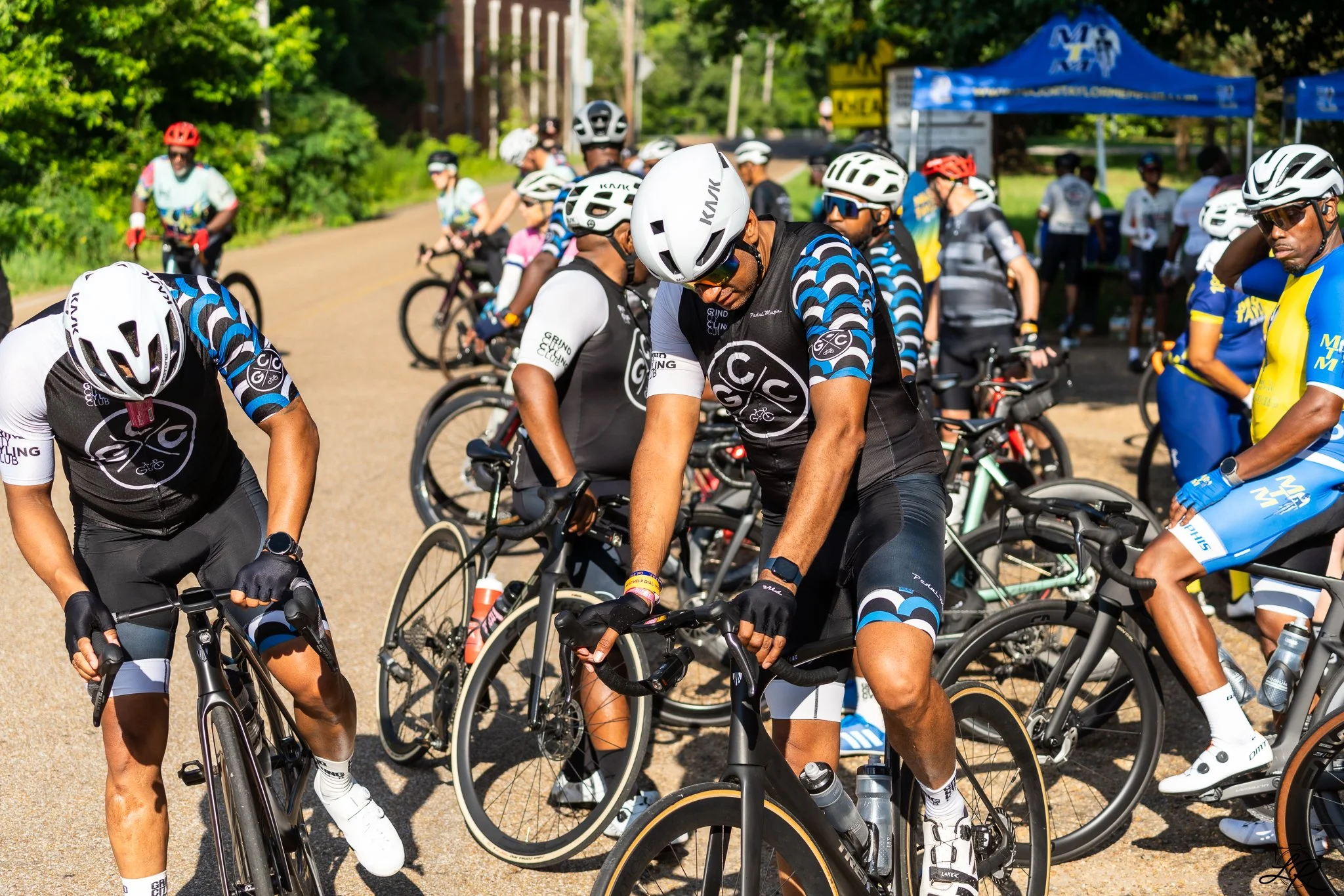 A group of cyclists wearing helmets and cycling gear gathered outdoors with trees and tents in the background, preparing for a ride or race.