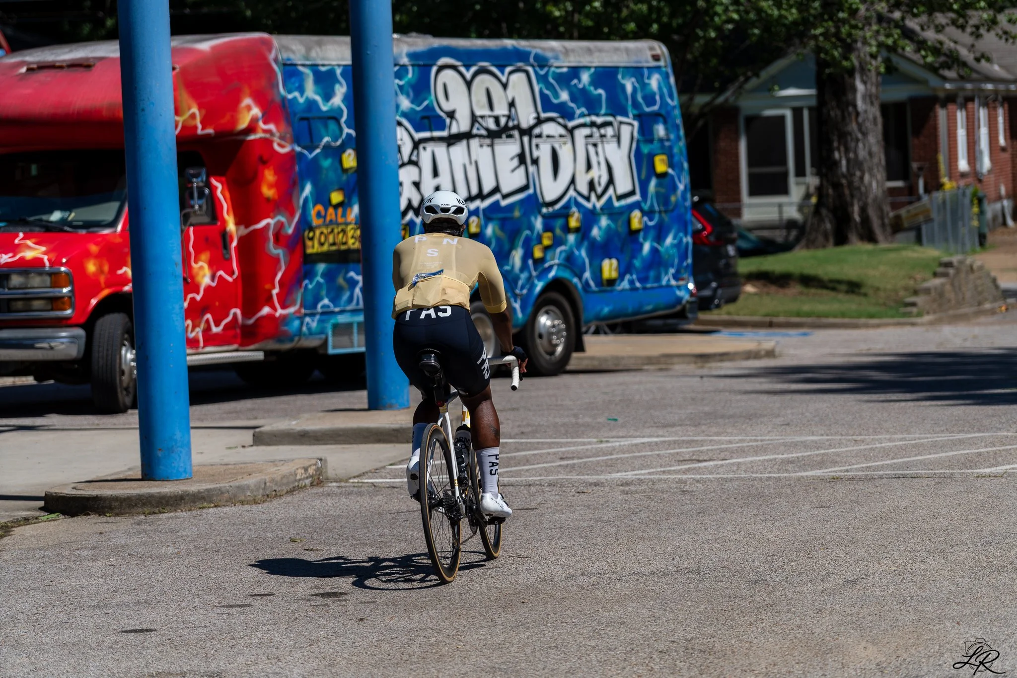 A cyclist riding a bicycle on a city street near a graffiti-covered bus and parked cars. The cyclist is wearing a helmet and sports attire, and the street has blue poles and trees in the background.