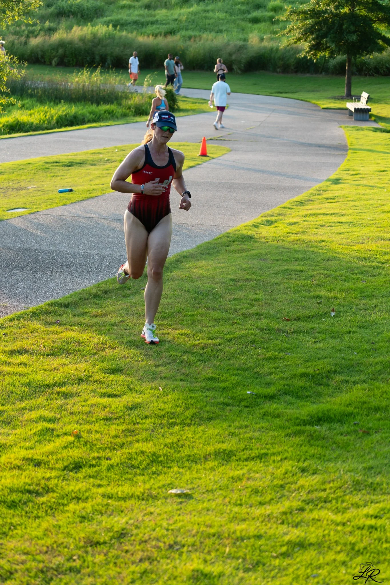 A woman running outdoors on a paved pathway in a park, with several people walking in the background, green grass, trees, and a bench visible.