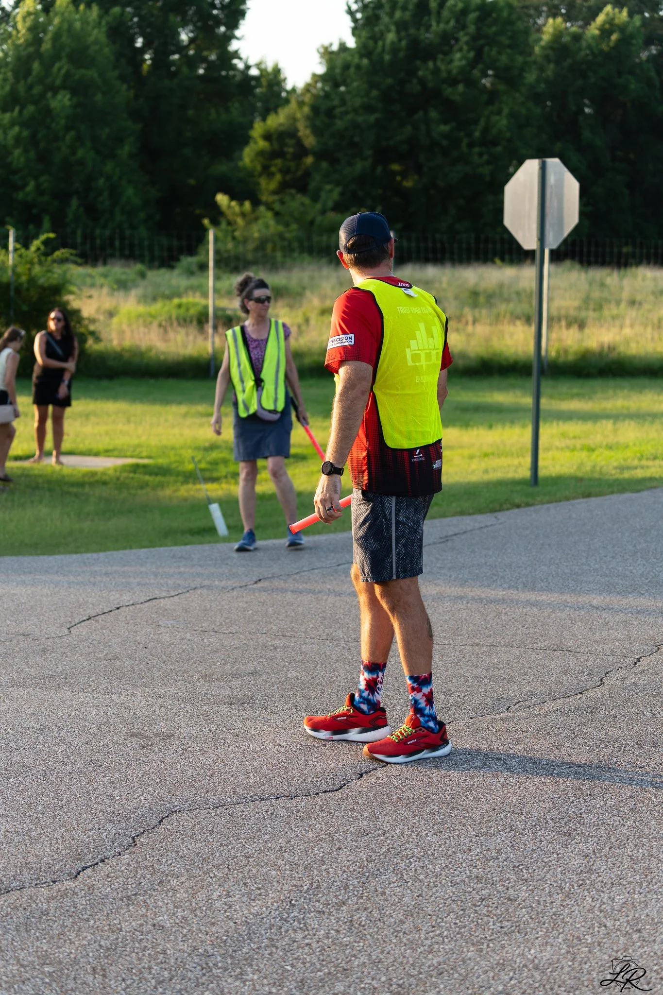 A man in athletic clothing holding an orange baton standing on a cracked asphalt road, with two women and a girl in the background, some trees, and a stop sign.