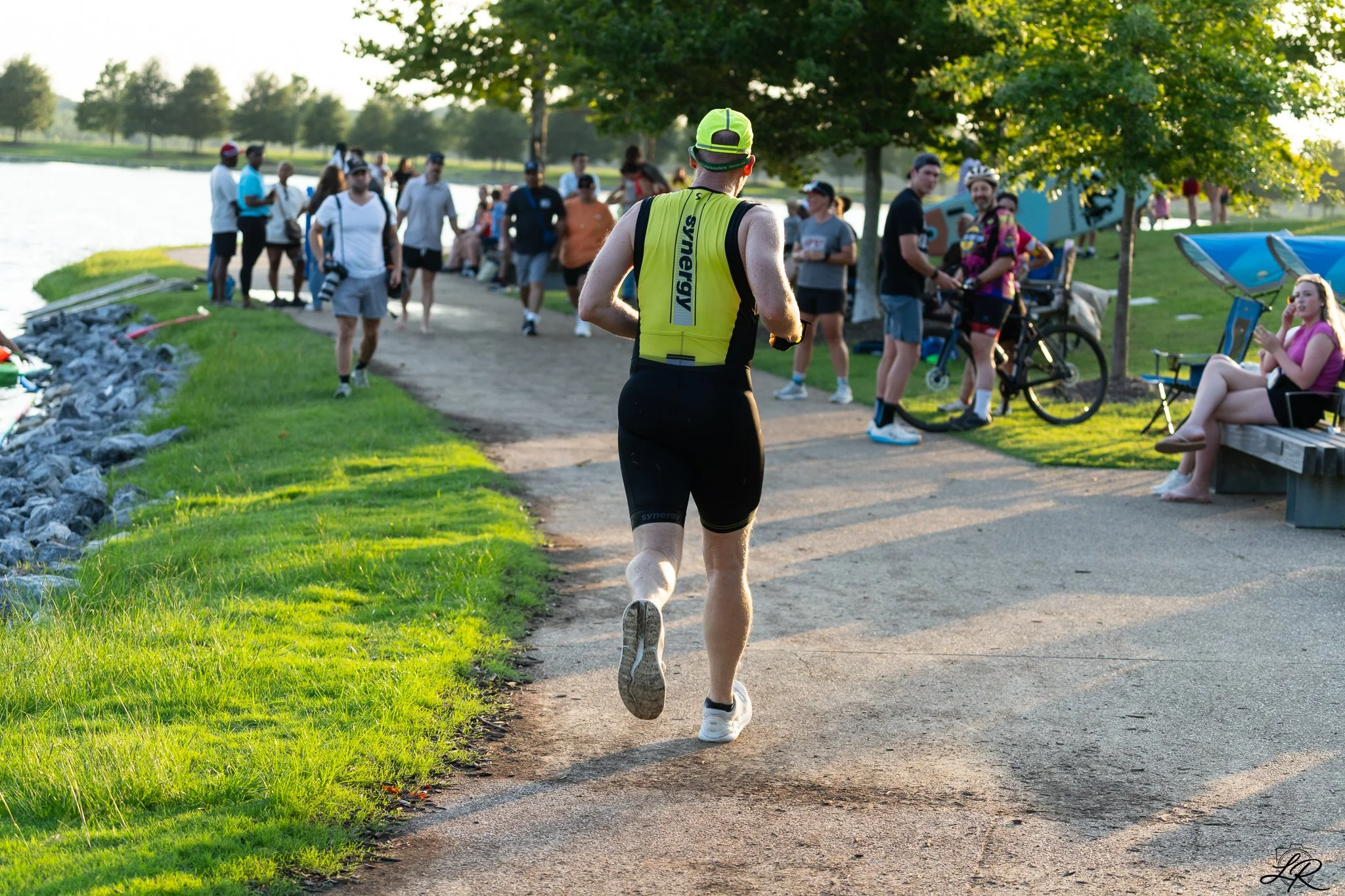 A man running on a path near a lake during a daytime outdoor event, with a crowd of people watching and taking photos nearby, surrounded by green trees and grass.