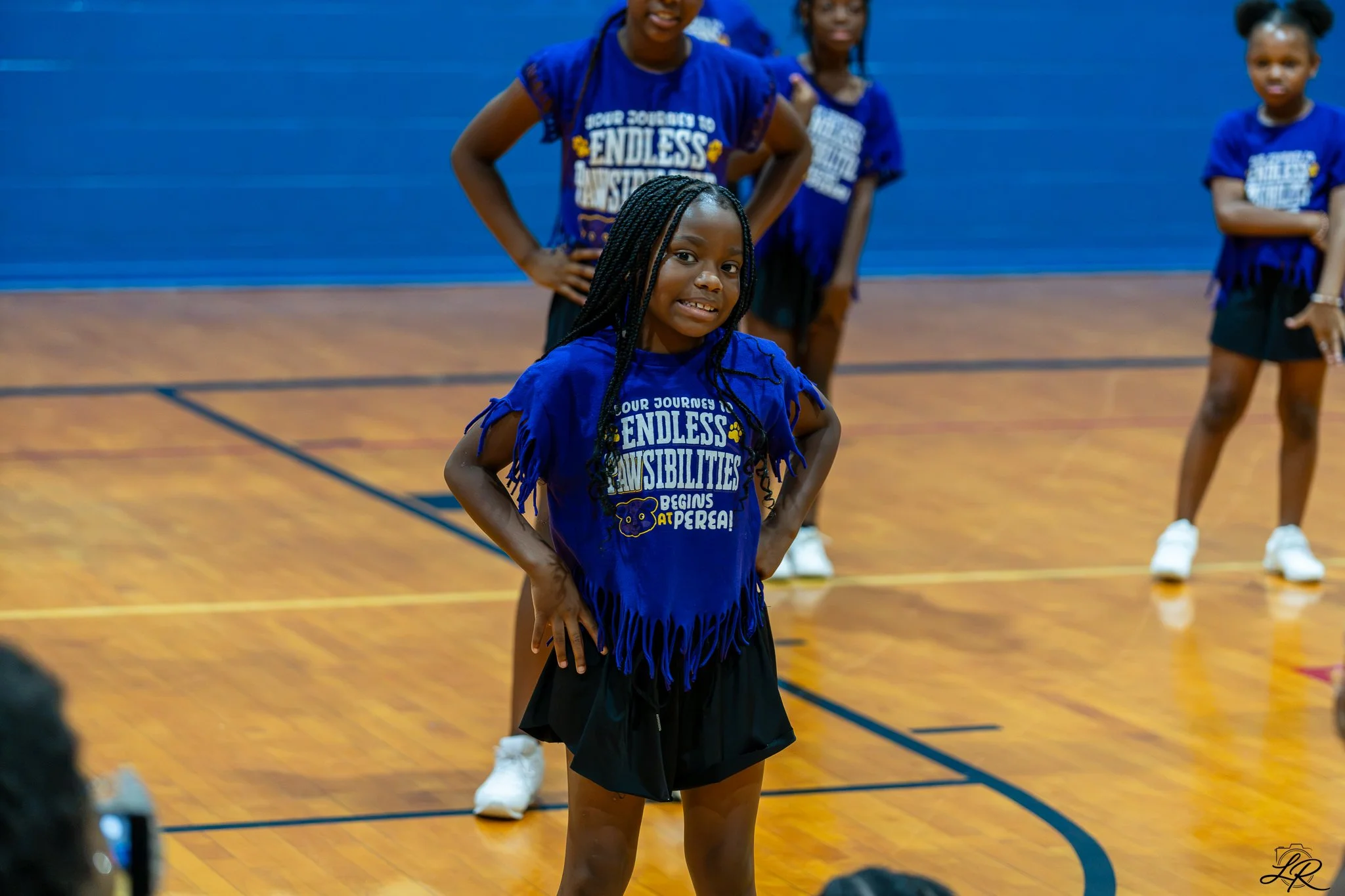 Young girl in black skirt and blue fringed shirt standing in a gymnasium, surrounded by other children wearing similar blue shirts.