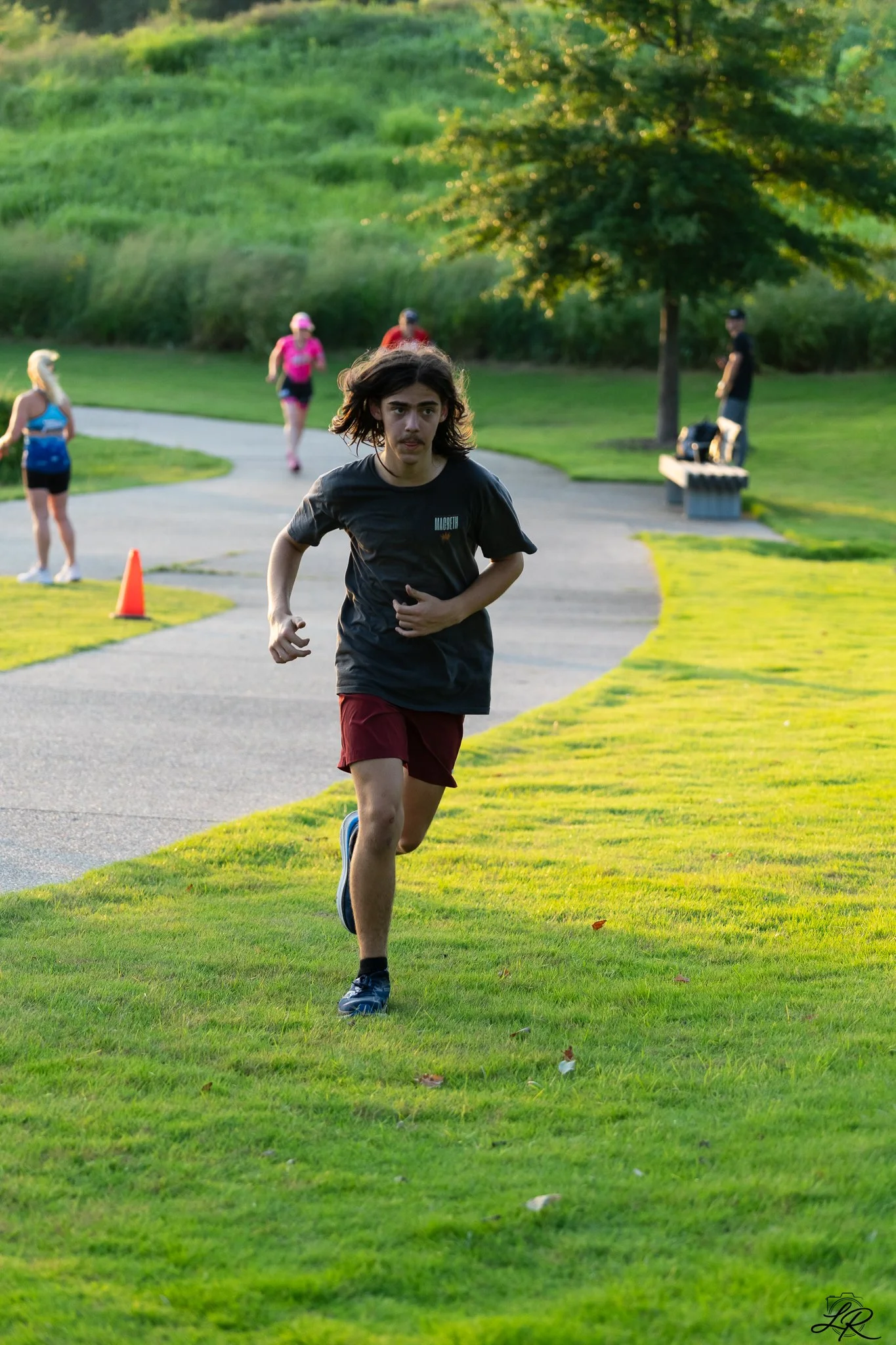 A young man with long hair and a mustache running on a grassy park path during daylight. Other people are walking or jogging in the background, with some wearing athletic clothing. There are trees and a bench in the park.