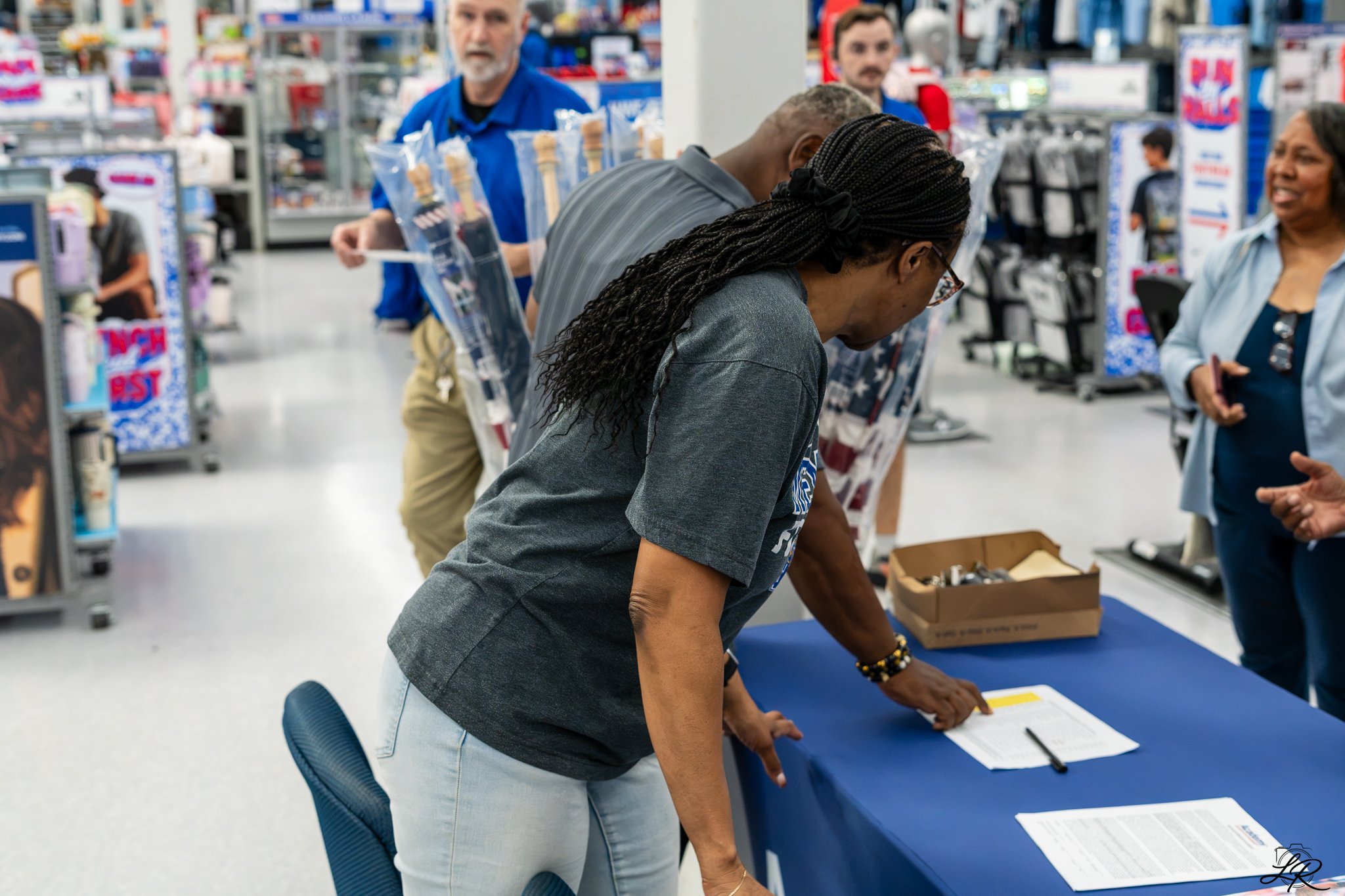 People standing in line at a store checkout counter, with a woman leaning over to pick an item and a man with gray hair and beard in blue uniform in background.