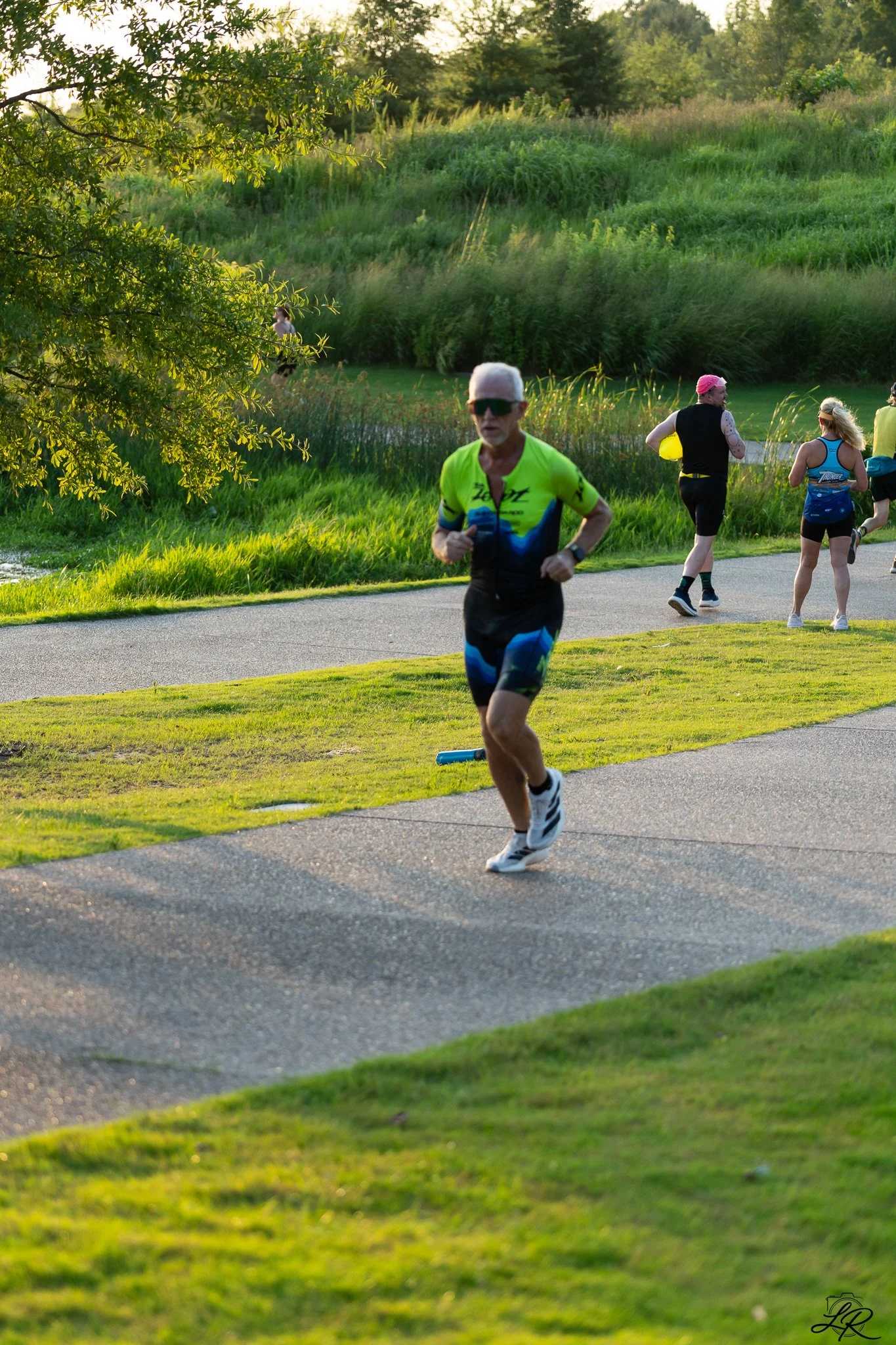 A group of runners participating in a race along a paved trail in a park with lush green grass, trees, and shrubs during a sunny late afternoon.