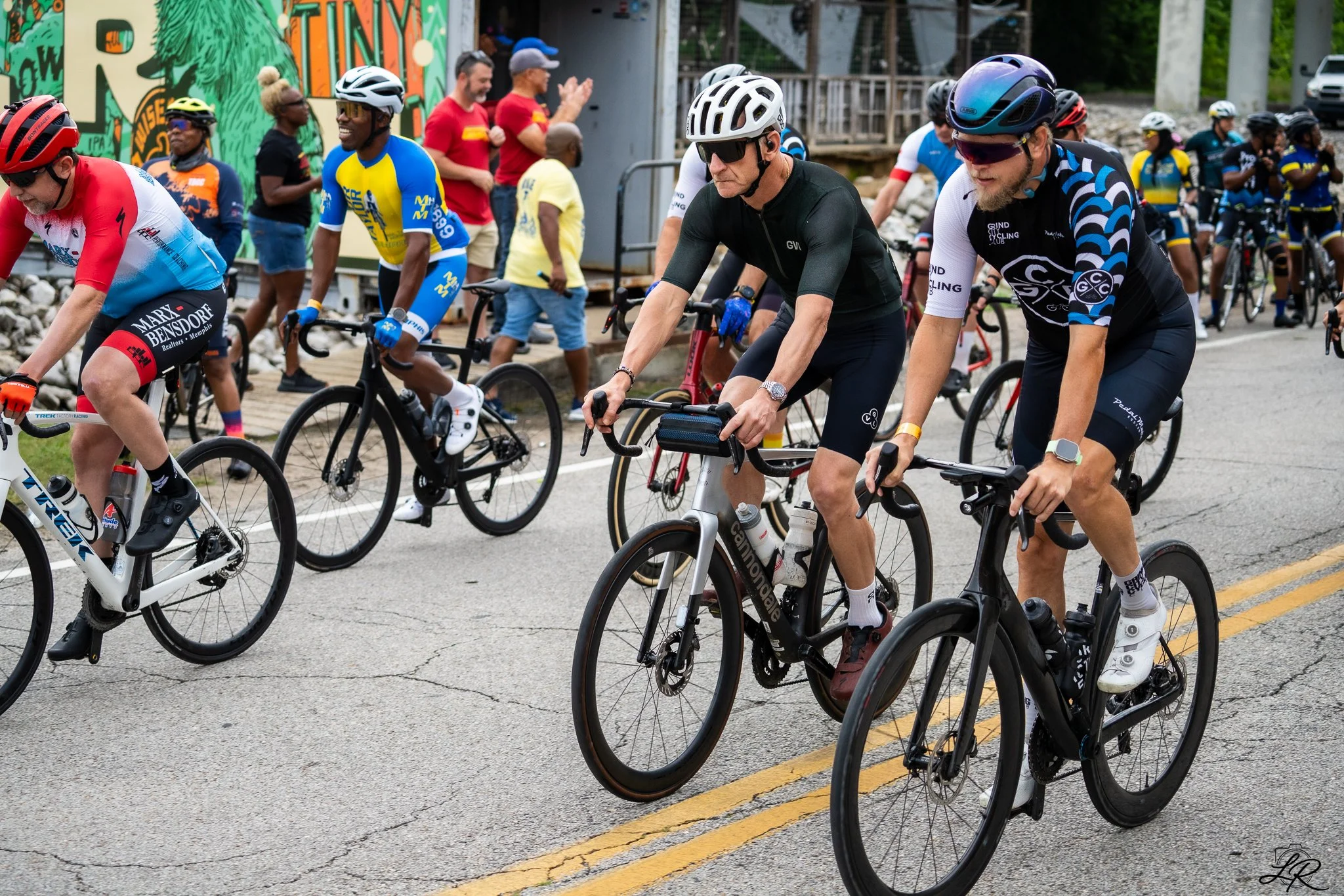 A group of cyclists riding in a race, wearing helmets and colorful athletic clothing, with spectators along the street and a vibrant mural in the background.