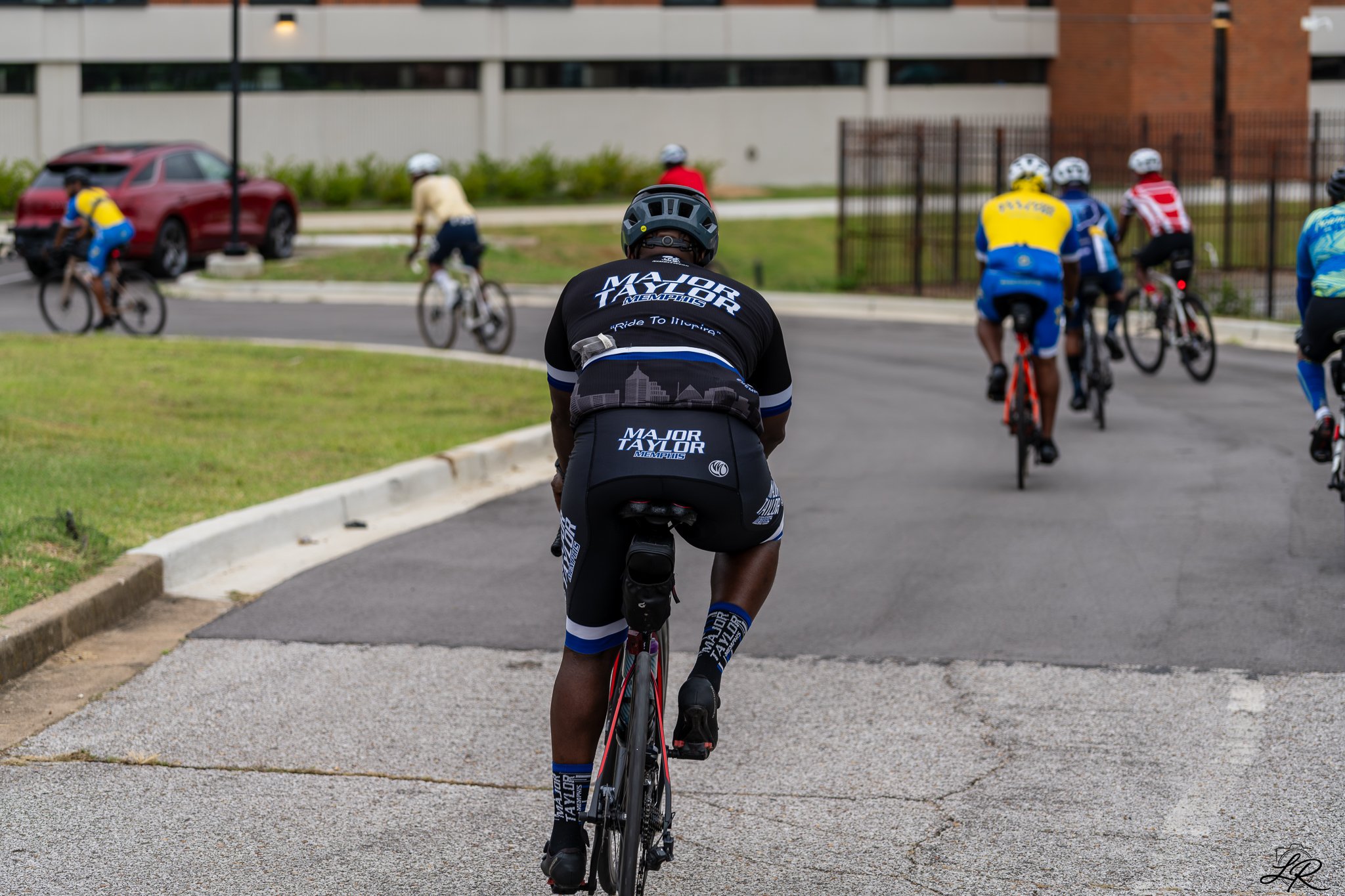 A group of cyclists riding on a paved road, with one cyclist in the foreground wearing a black helmet and a black jersey that says 'Major Taylor Memphis', and other cyclists in colorful jerseys in the background.