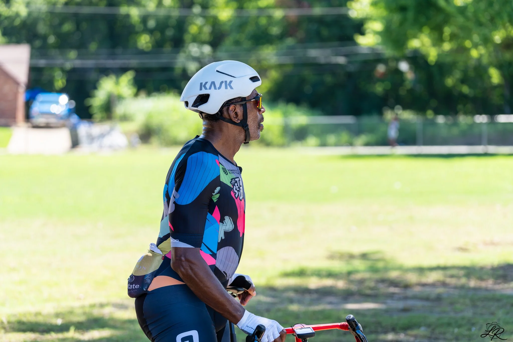 A man wearing a white helmet with the word "KASK" on it, sunglasses, a colorful cycling jersey, and blue cycling shorts, standing outdoors on a grassy area with a bicycle, in front of a blurred background of green trees and power lines.