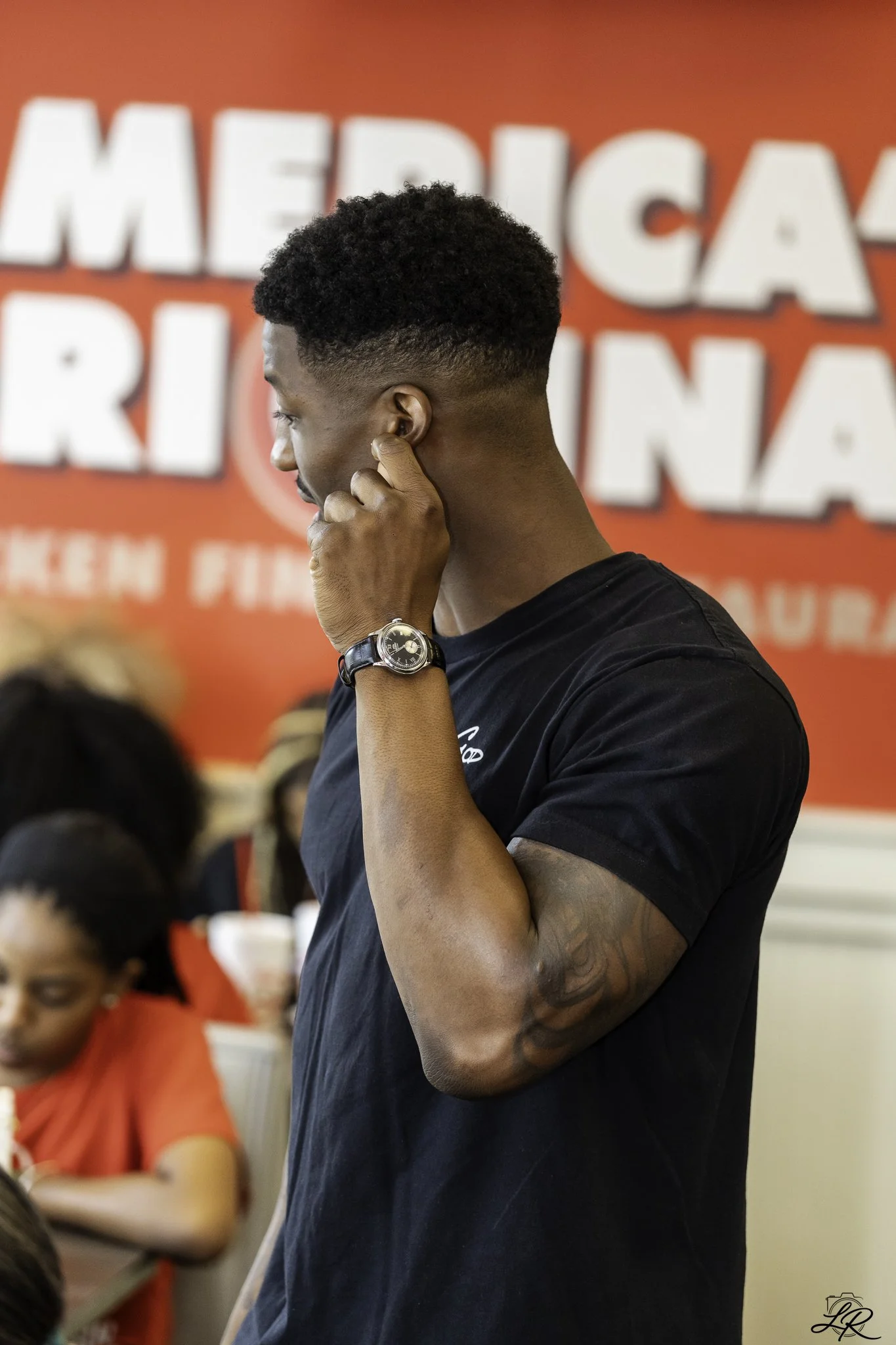 A side view of a young man with short curly hair and a fade haircut, wearing a black T-shirt and a wristwatch, standing in front of a red and white background with partially visible large text.