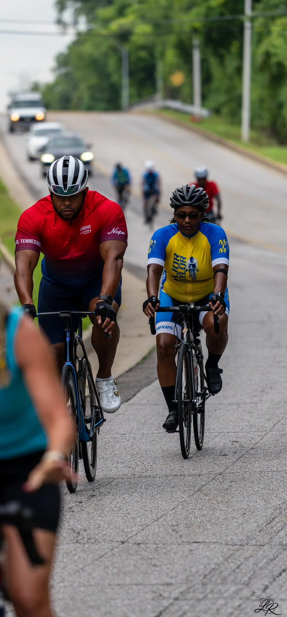 Two cyclists riding on a suburban road with cars and greenery in the background, wearing helmets and cycling jerseys.