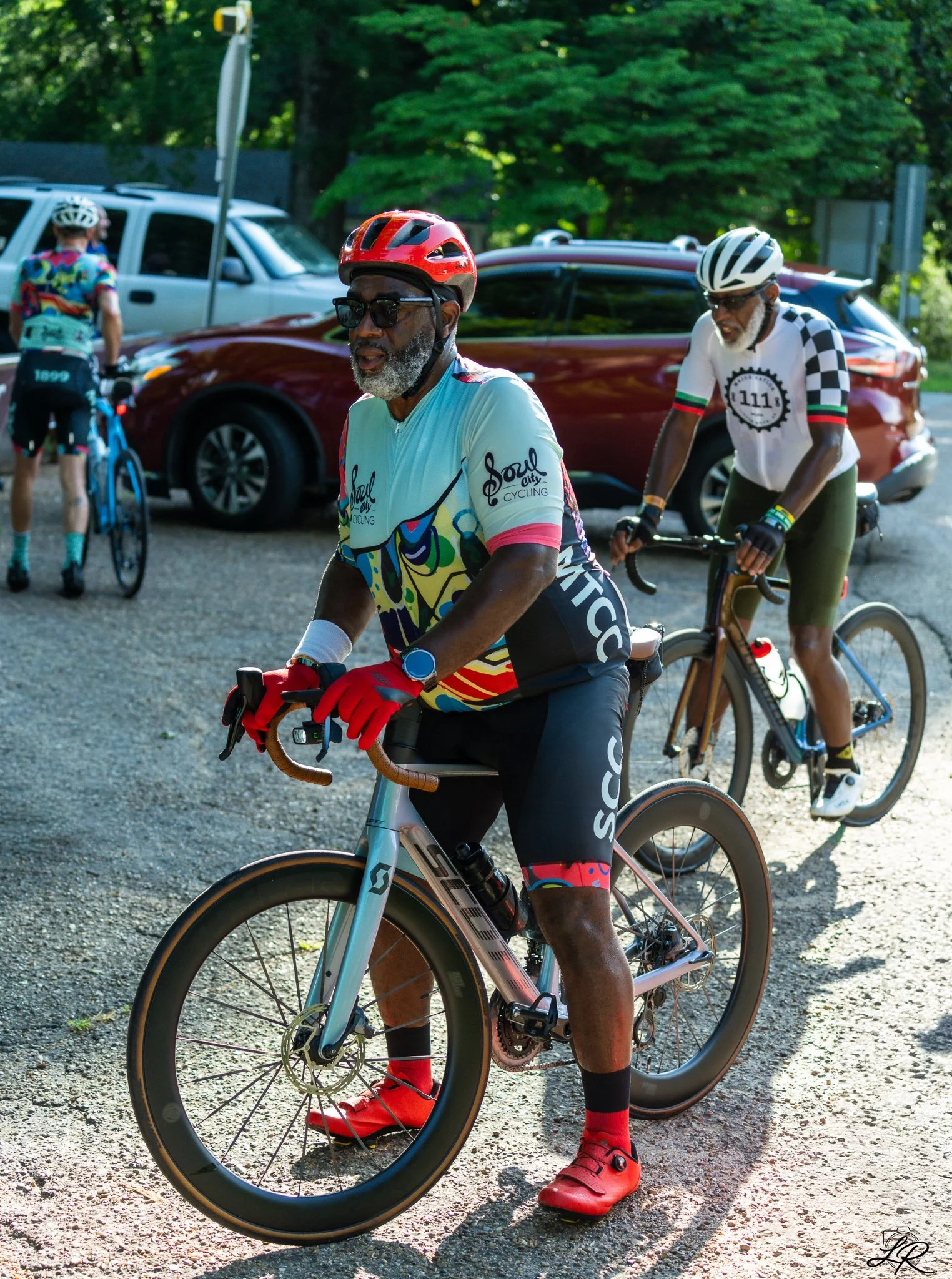 Two cyclists riding their bikes on a gravel road, with a man in colorful cycling attire in the foreground, wearing a red helmet, sunglasses, and red shoes. A man in a white and checkered cycling jersey stands behind him. Two cars are parked in the ba