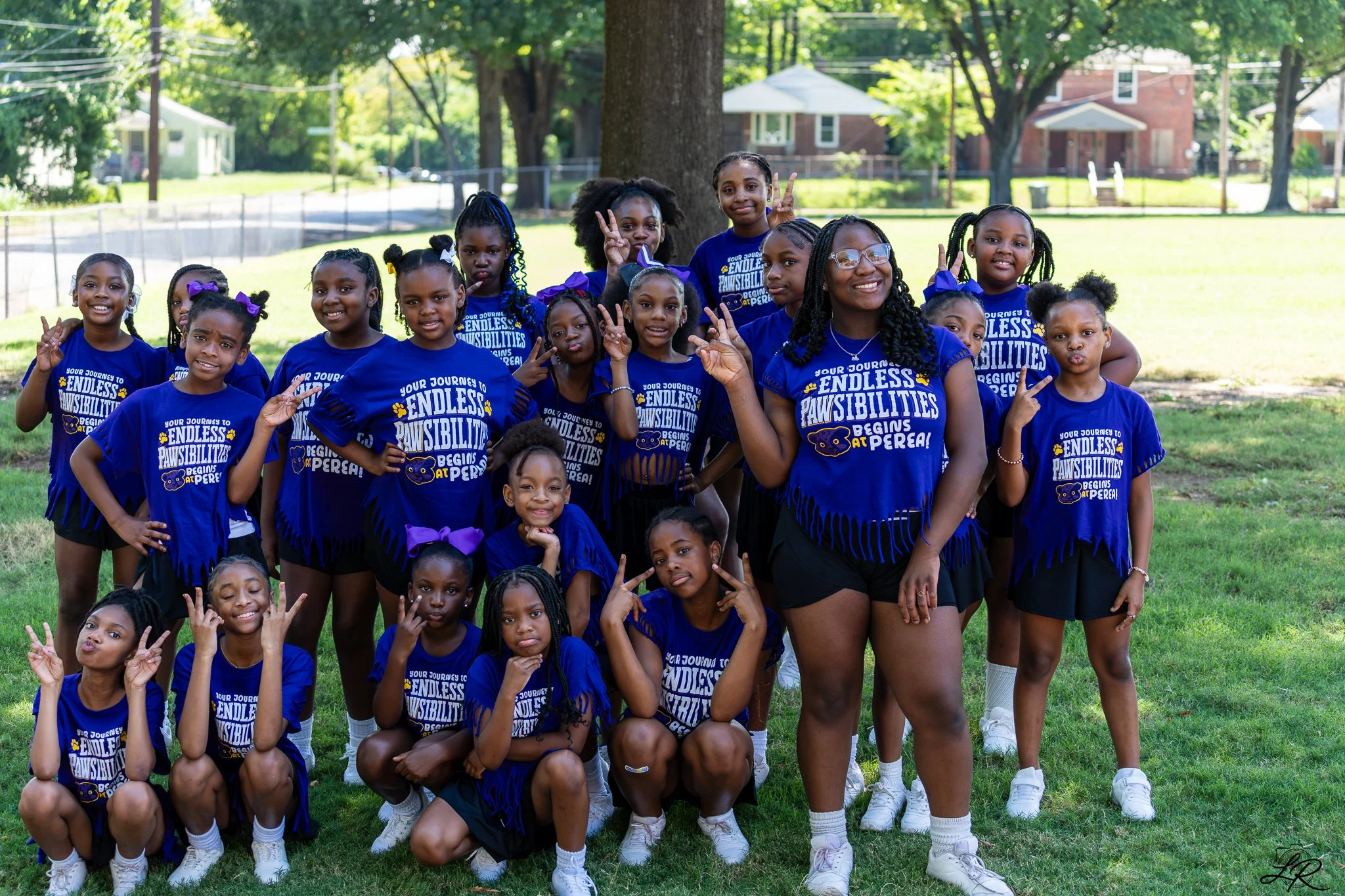 A group of young girls and a woman, all wearing matching blue T-shirts, posing outdoors in a park with trees and houses in the background. The shirts have the text 'Your Journey to Endless Pawsibilities Begins at Perea!'.