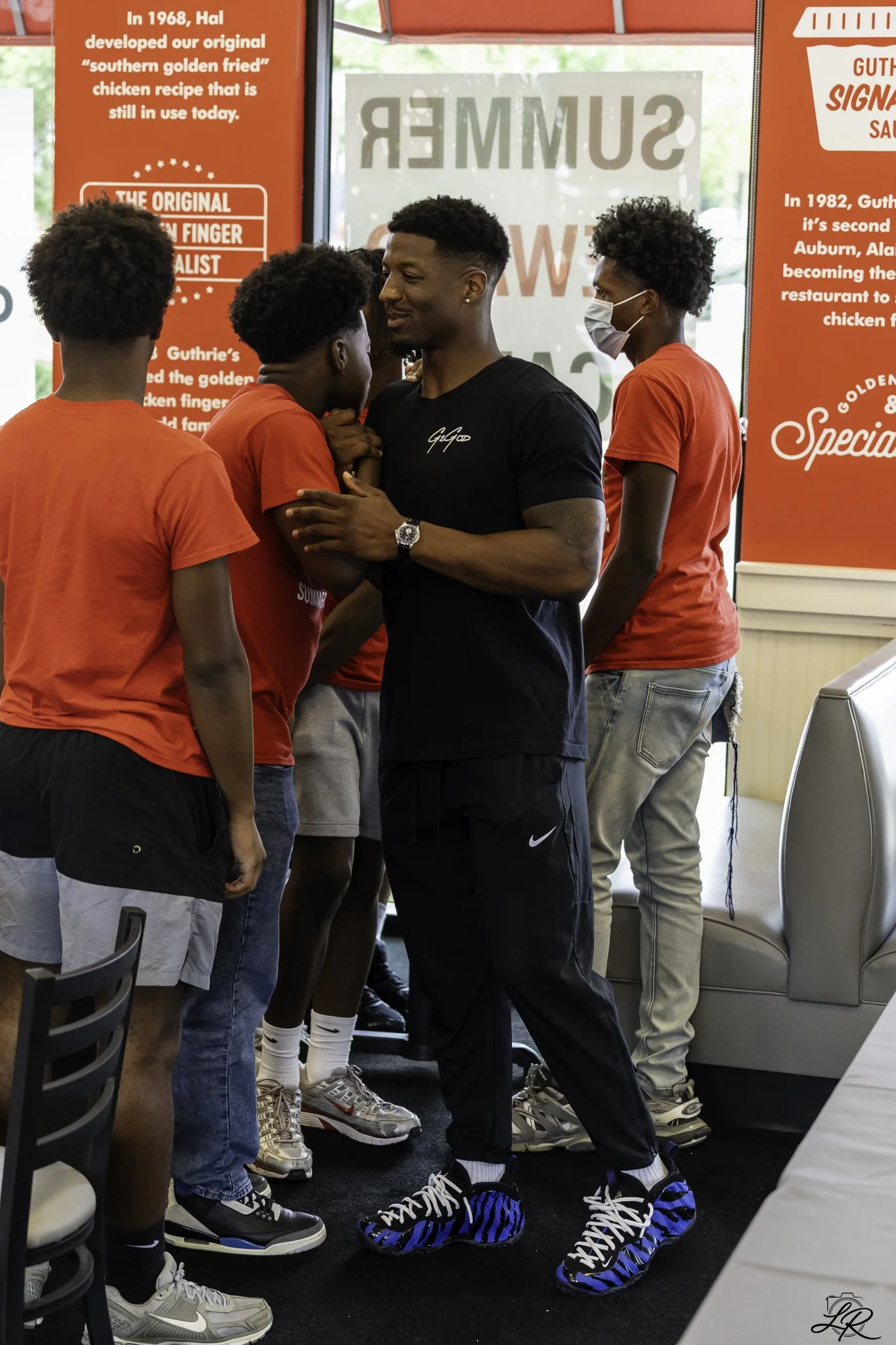 A group of young men in casual sportswear, some wearing masks, standing indoors. Two of them are engaging in a close, friendly conversation, with one touching the other's face. The background shows red walls with text about historical restaurant mile