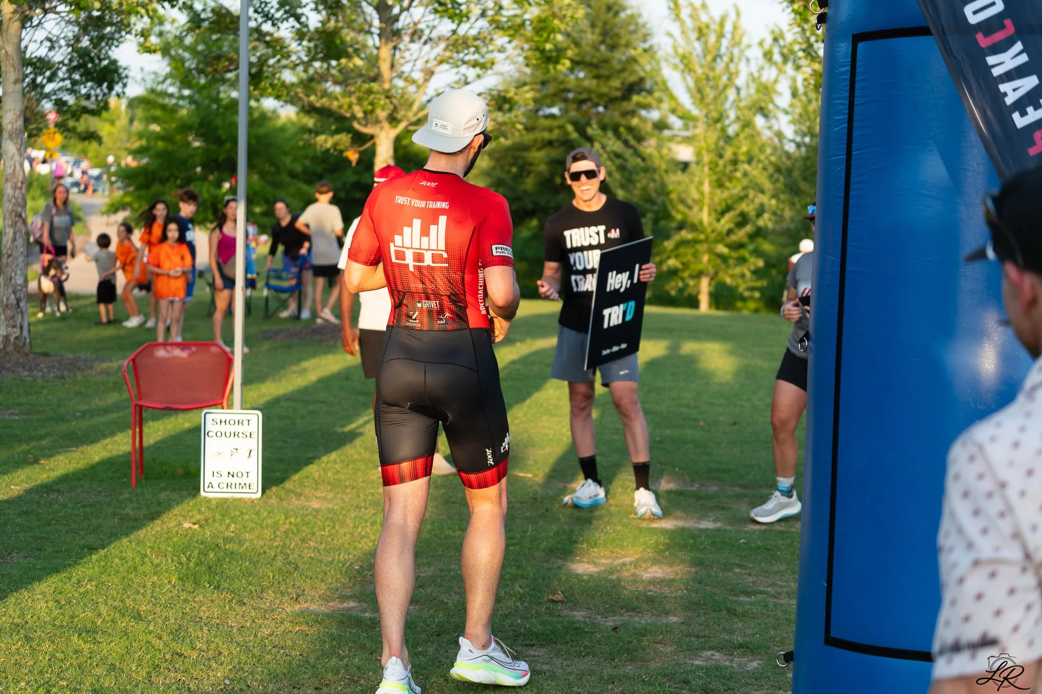 A man wearing a red cycling jersey and black cycling shorts is talking to a woman at a running event. The background shows other participants and spectators, along with a white sign that says 'Short Course - Is Not a Crime.' Trees and greenery are vi
