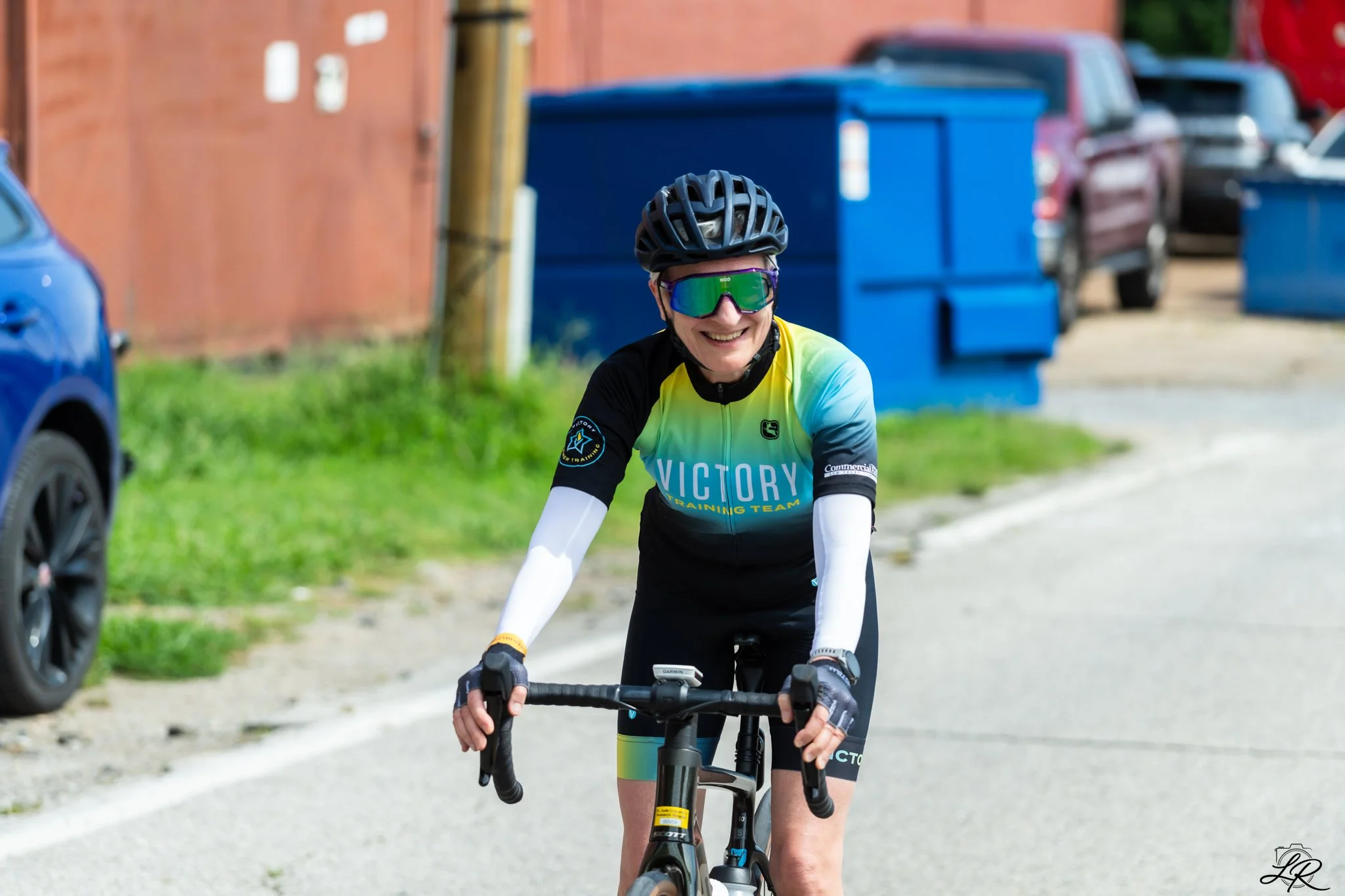 A woman wearing a black helmet and reflective sunglasses riding a bicycle on a street, smiling. She is dressed in a blue, green, and black cycling jersey with the words "Victory" and "Training Team" on it. In the background, there are parked cars and