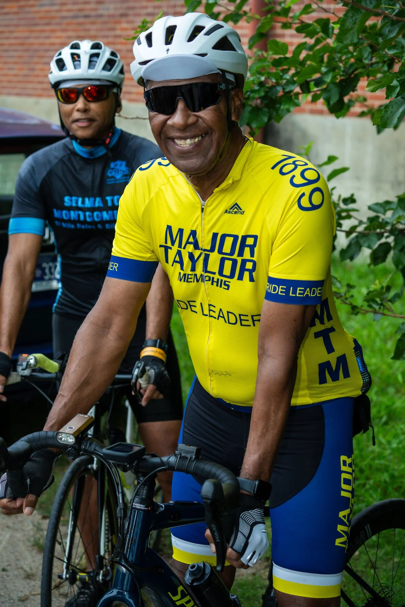 Two men with helmets and sunglasses, smiling, wearing cycling gear, standing with mountain bikes on a trail next to green bushes and a brick wall.