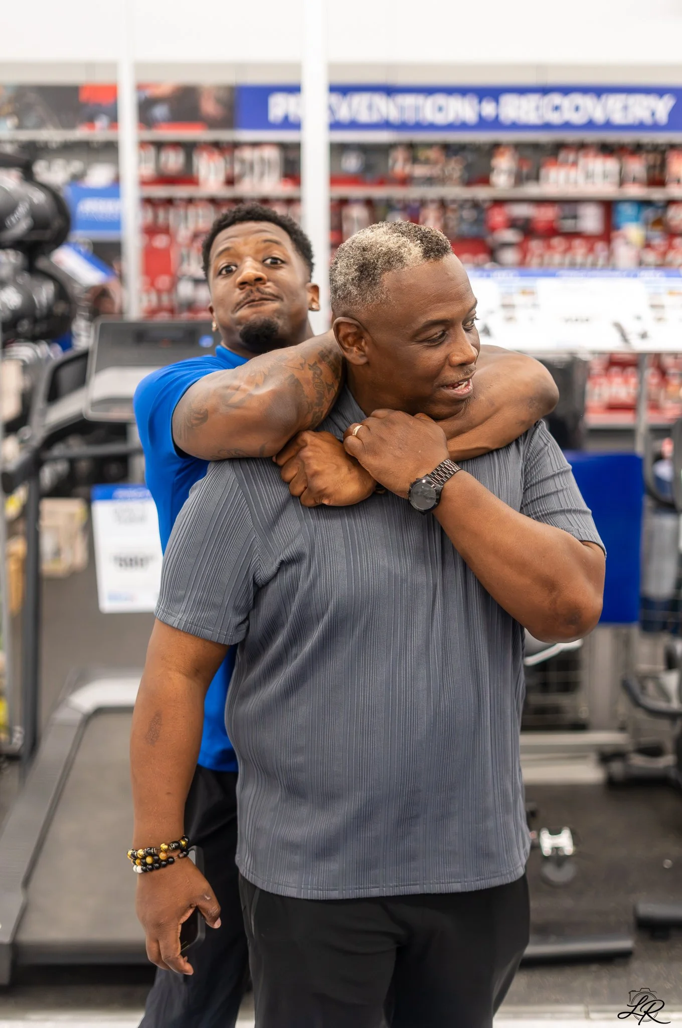 Two men in a store, one with blond hair and gray hair, the other with curly black hair, engaged in a playful wrestling pose, with supermarket shelves in the background.