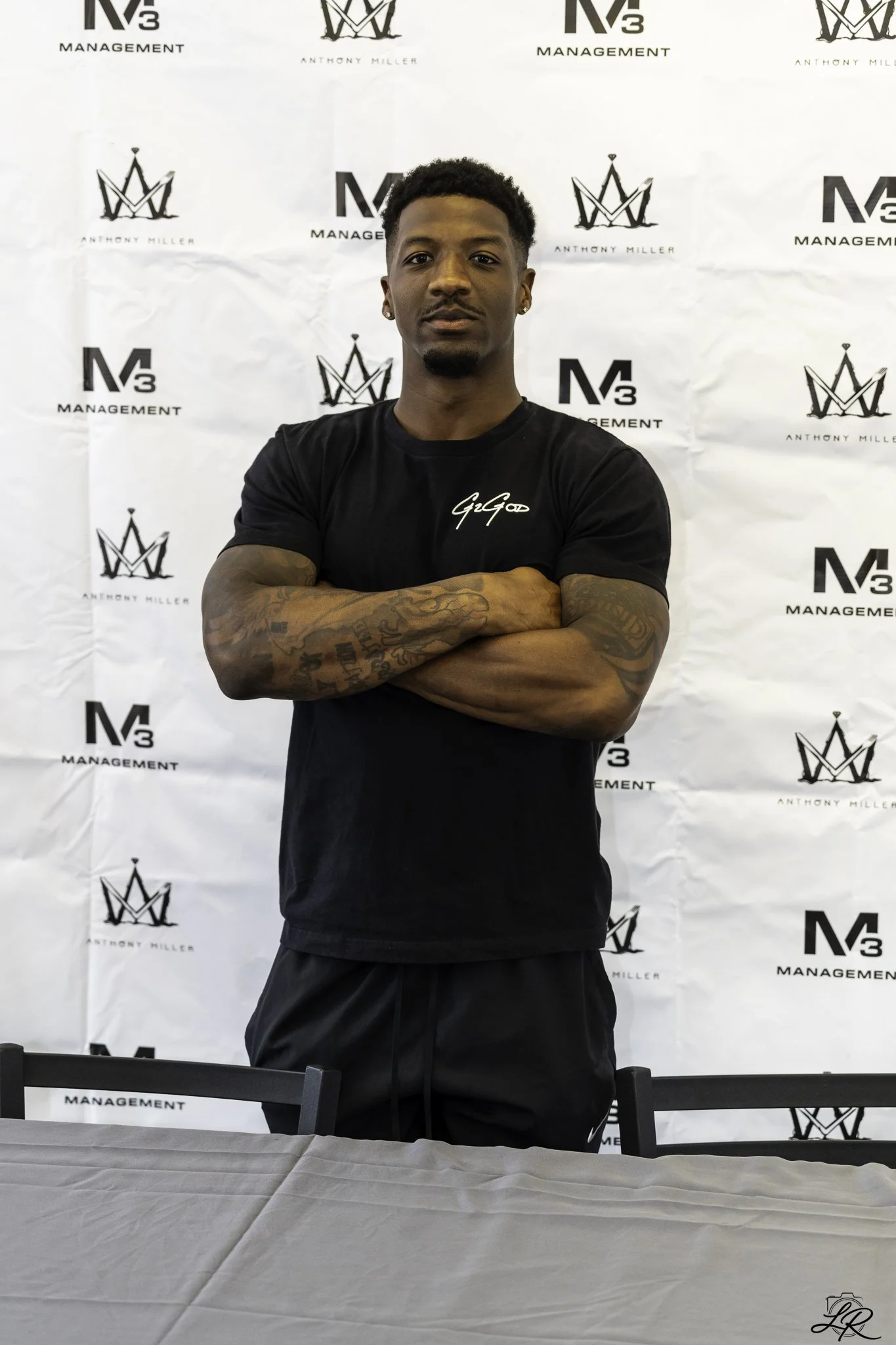 A young man with short curly hair and earrings standing with arms crossed in front of a backdrop with logos that say 'M3 Management' and 'Anthony Miller'. He is wearing a black t-shirt and black shorts, tattoos visible on his arm.