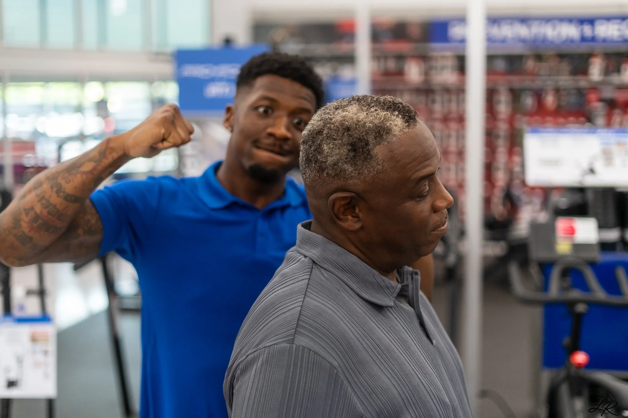 A young man with tattoos on his right arm is seen flexing his bicep while standing behind an older man with gray curly hair and a gray striped shirt, in a store aisle.
