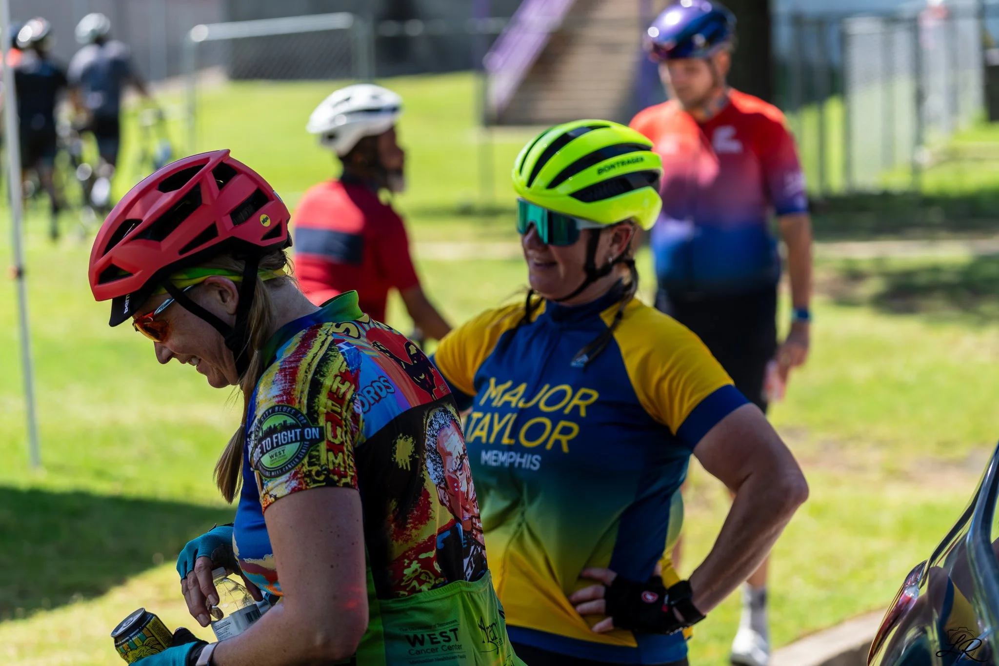 A group of cyclists wearing helmets and colorful jerseys at an outdoor event, smiling and conversing, with a grassy area, fence, and a few people in the background.