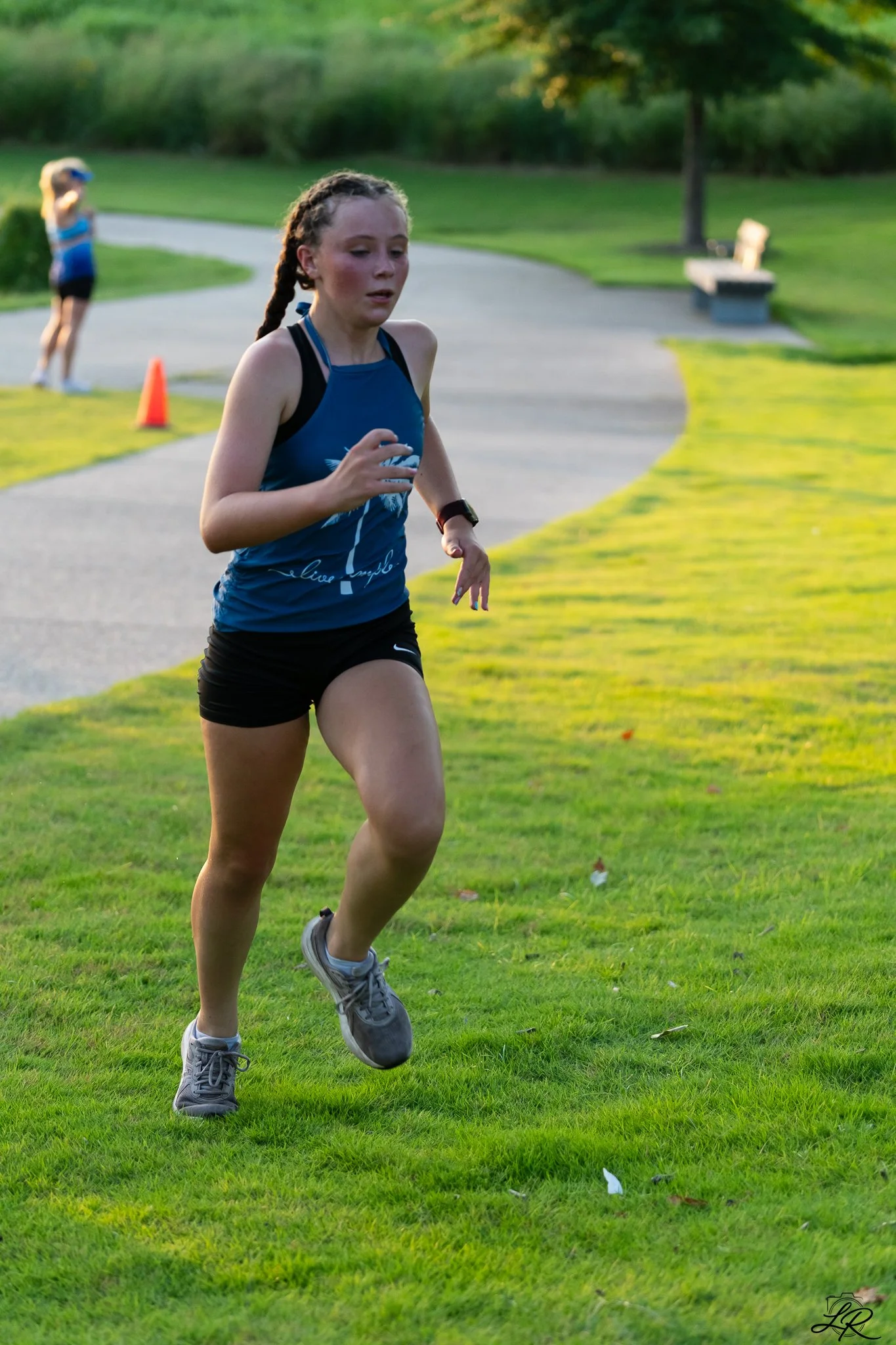 A young girl with braids running on grass in a park during daylight, with another girl in the background on a curved pathway, trees, a bench, and orange cone.