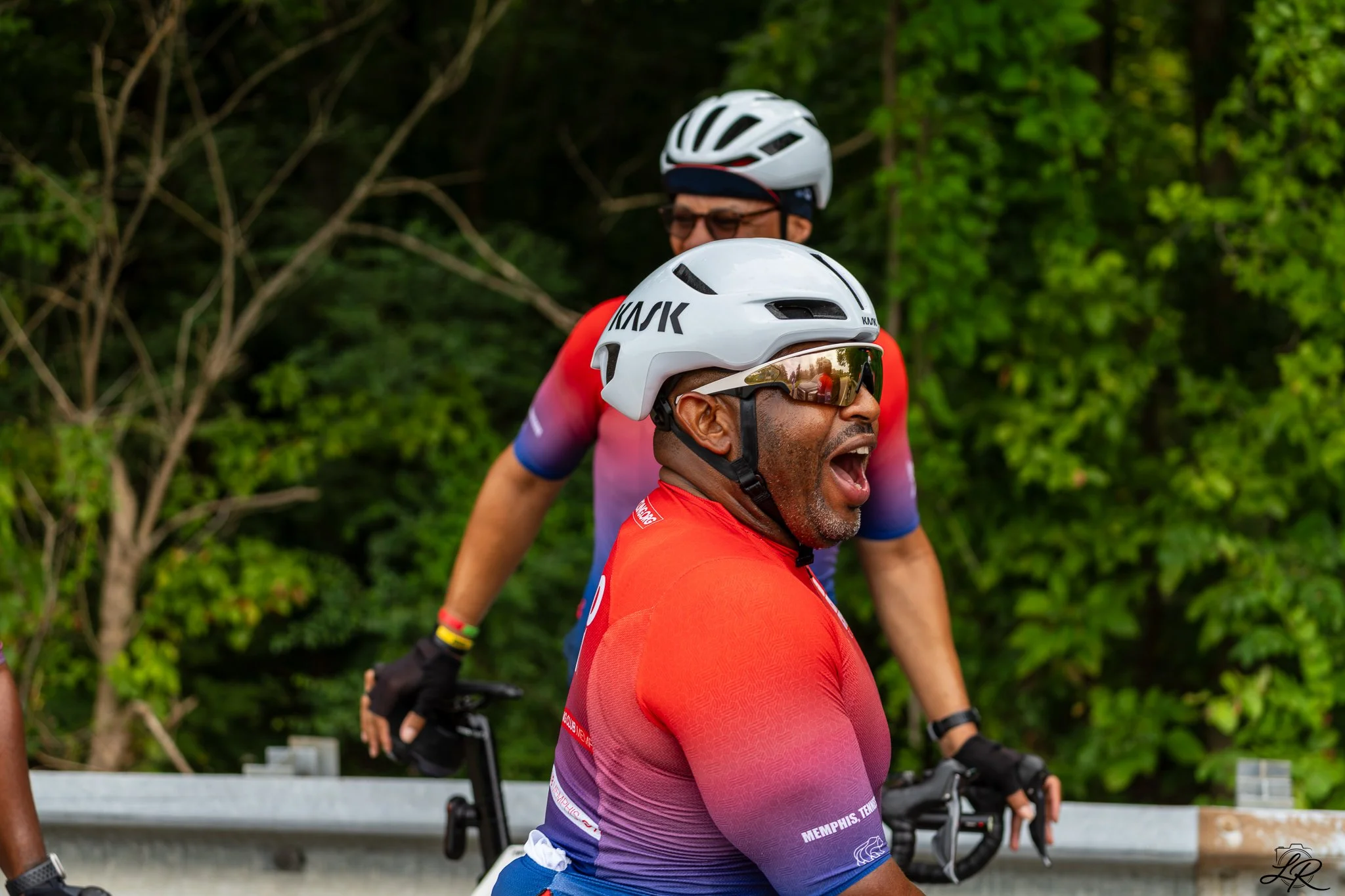 Two men wearing white bicycle helmets and cycling jerseys, one in red and the other in a multicolored gradient, are outdoors among green trees and shrubbery, riding bicycles and talking.