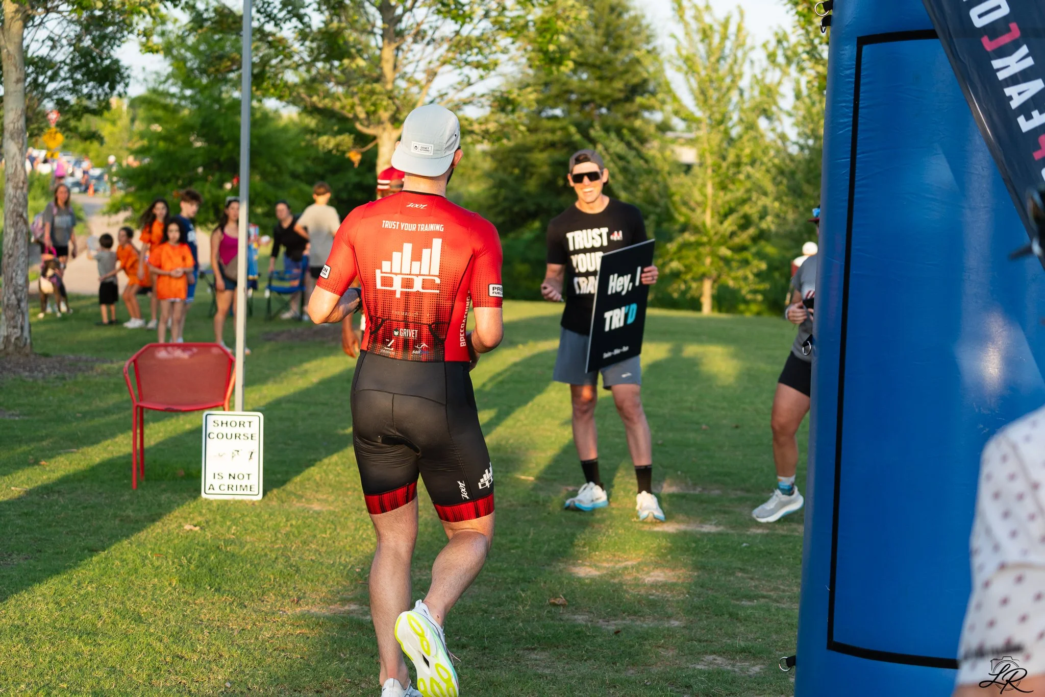 A male athlete wearing a gray cap, red cycling jersey, and black shorts running in a race event at an outdoor park. There are spectators in the background, some children and adults, and a person holding a sign that says "Hey, I TRID". A blue inflatab