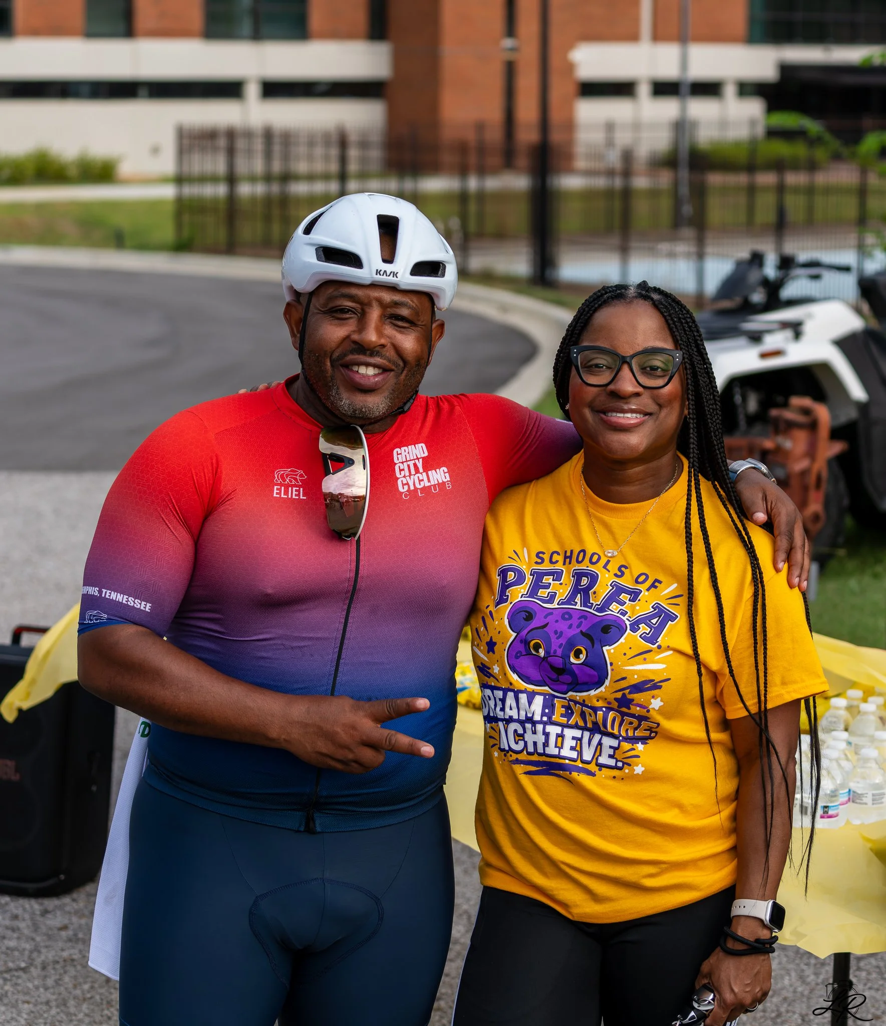 Two smiling African American adults, a man and a woman, posing outdoors. The man is wearing a white bike helmet and a red cycling jersey with words 'GRIND CITY CYCLING CLUB' and 'ELIEL.' The woman has long braided hair, glasses, and is wearing a brig