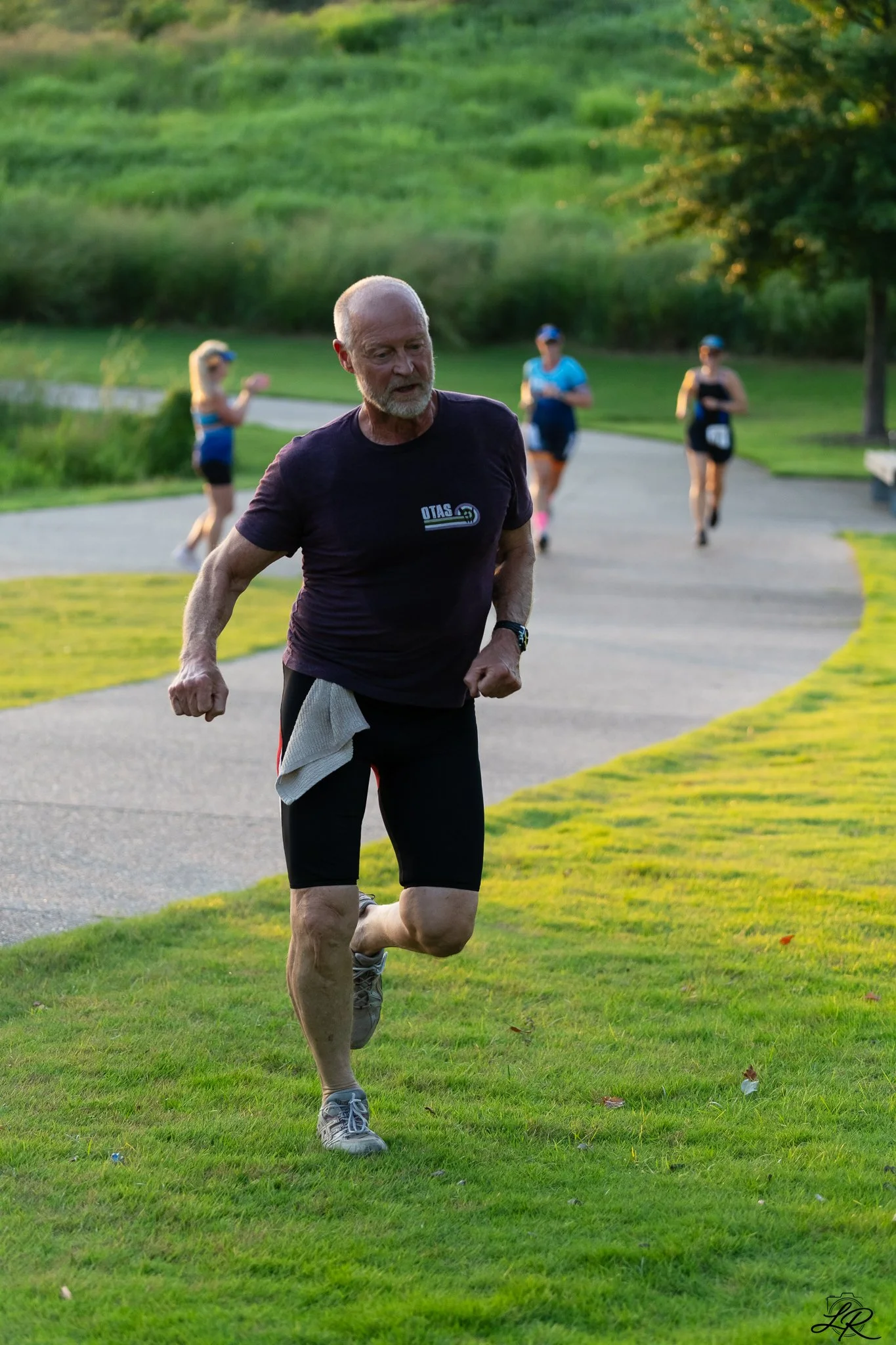 Older man running on a grassy park path, with three women in athletic clothing and caps jogging behind him, surrounded by green trees and a lush landscape.