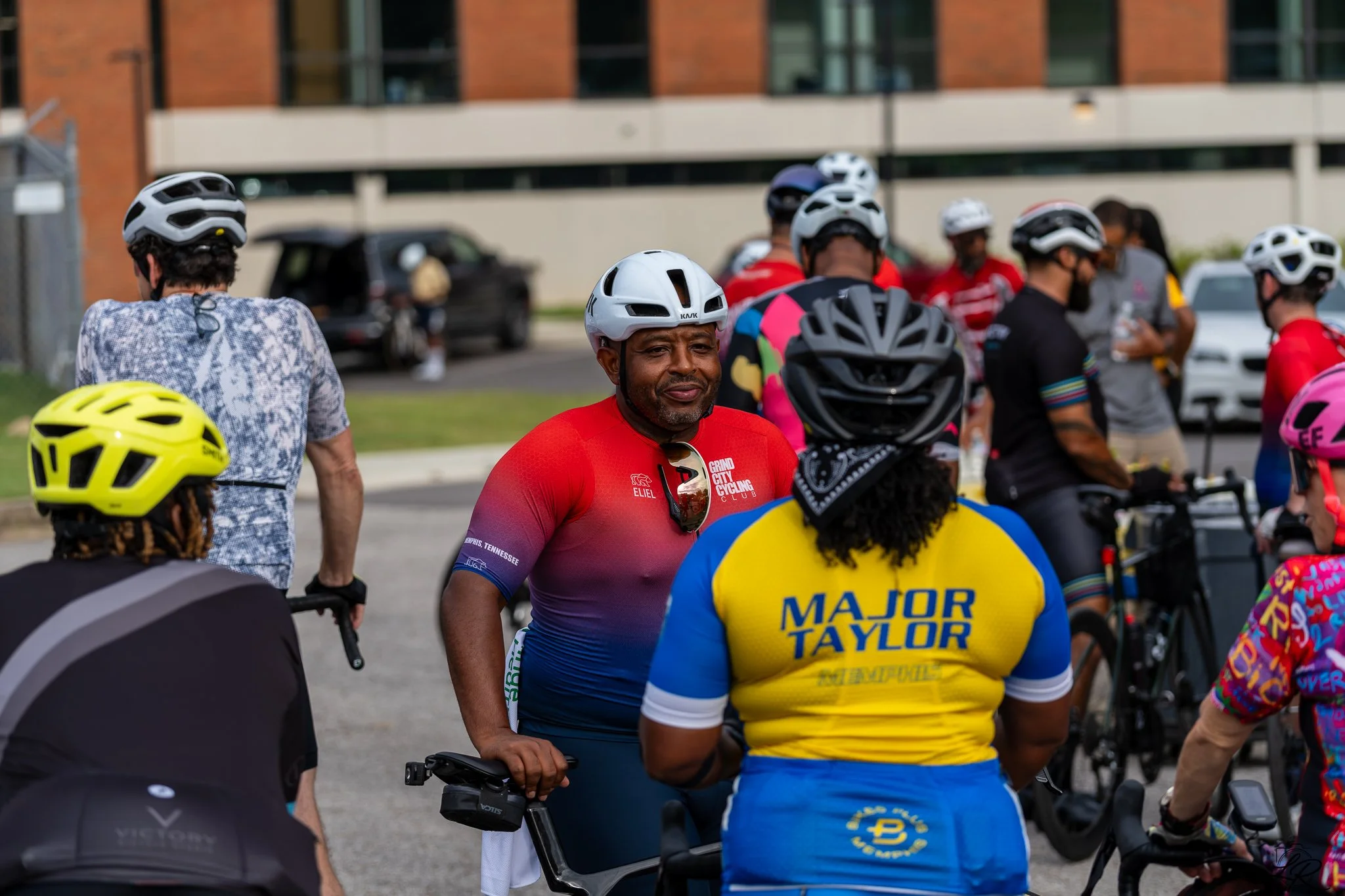 A group of cyclists gathered outdoors, wearing helmets and colorful cycling gear, with some engaging in conversation.