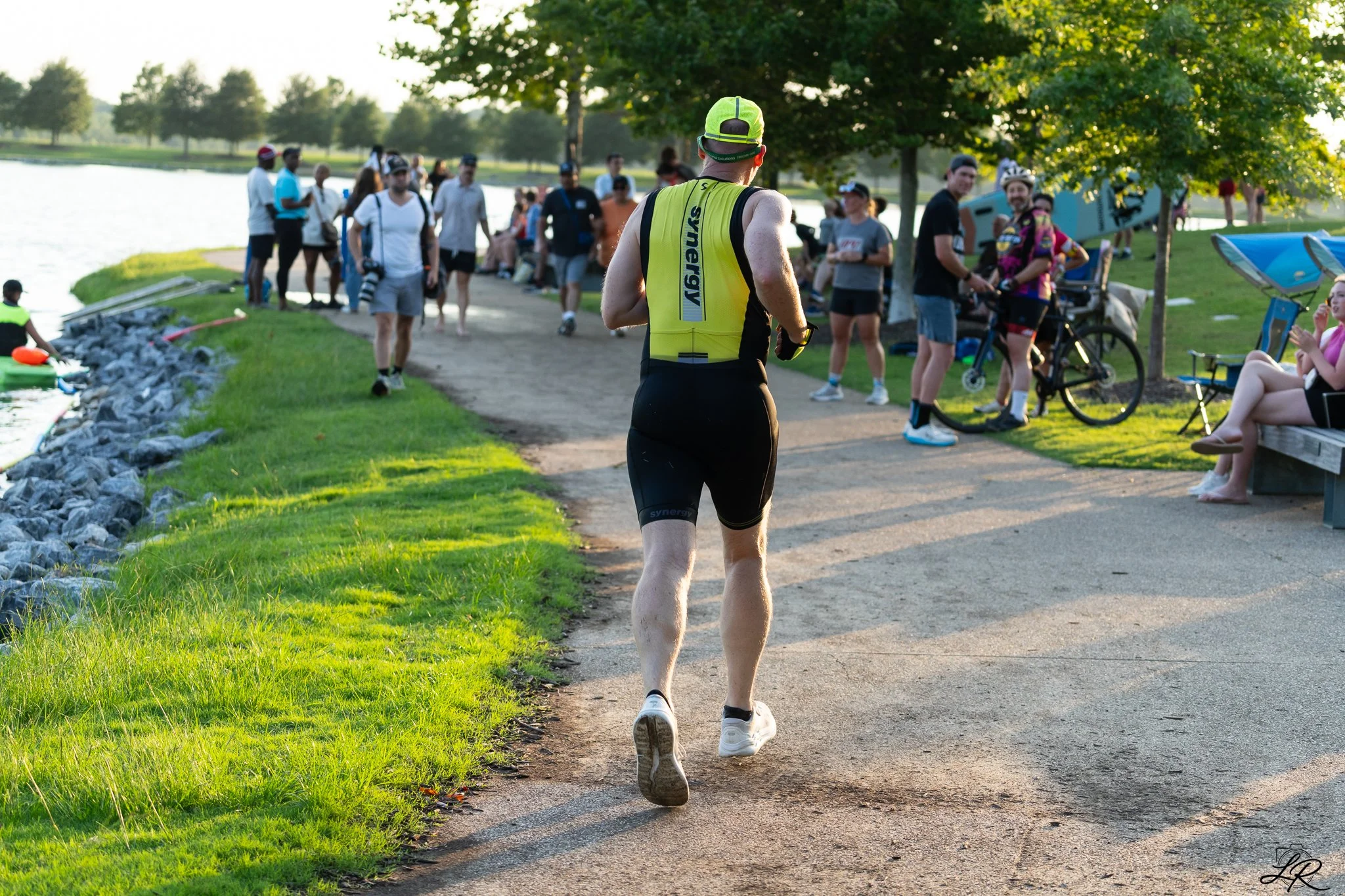A man running along a park pathway near a body of water with a crowd of people gathered, some sitting on benches and others standing, in the background during late afternoon or early evening.