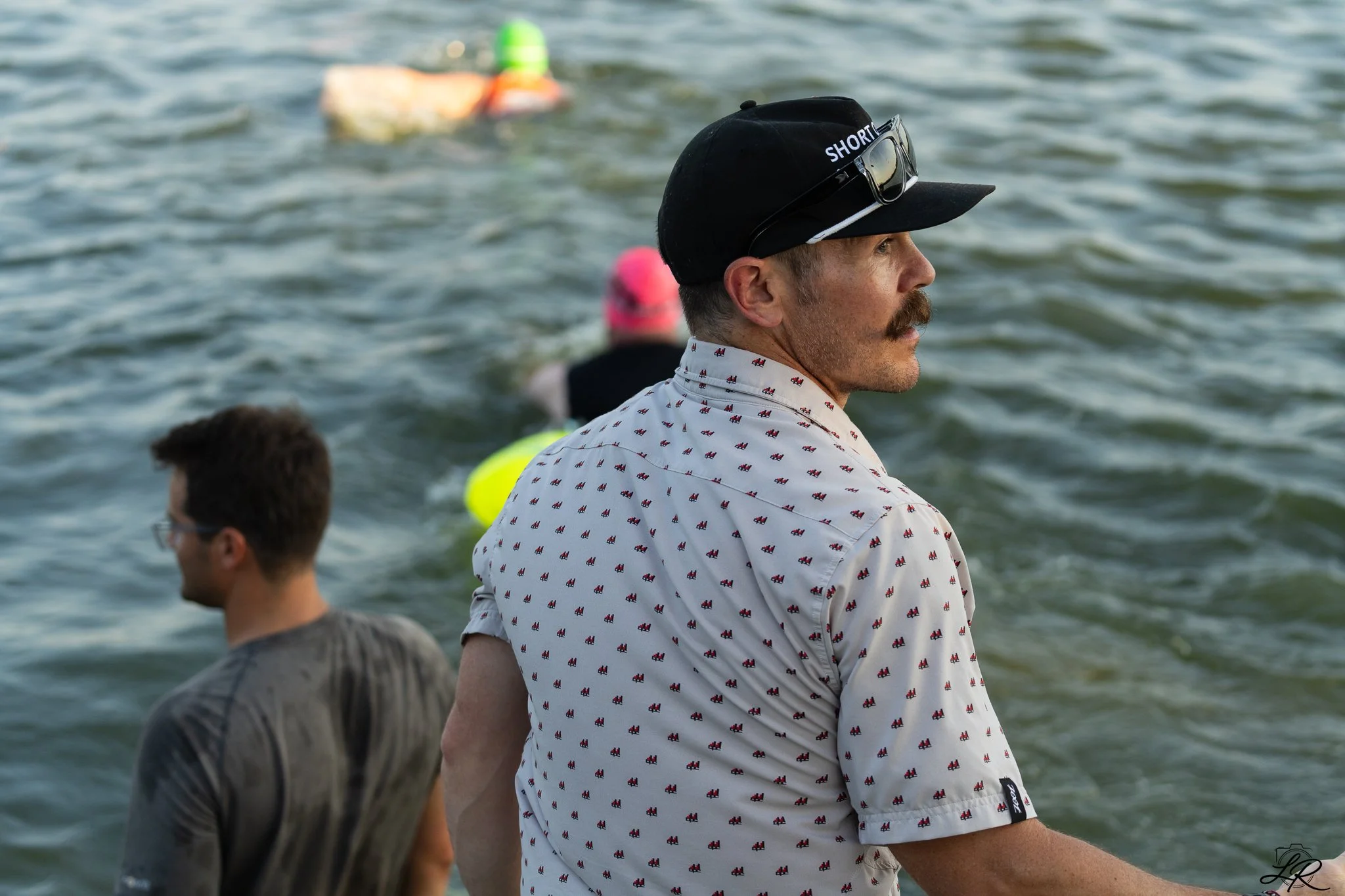 A man with a mustache wearing a black cap with 'SHORT' written on it, sunglasses, and a white shirt with small red and navy blue patterns, stands near water with other people around him.