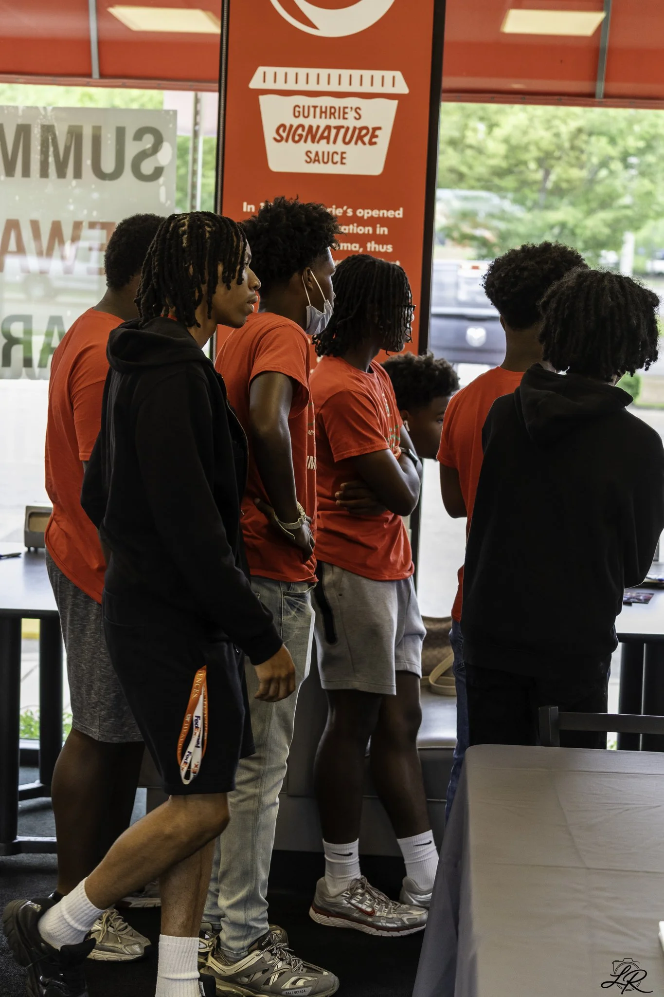 A group of young men in red and black clothing standing in line inside a fast food restaurant. They are wearing sneakers, and some are wearing masks, with a red sign advertising 'Guthrie's Signature Sauce' behind them.