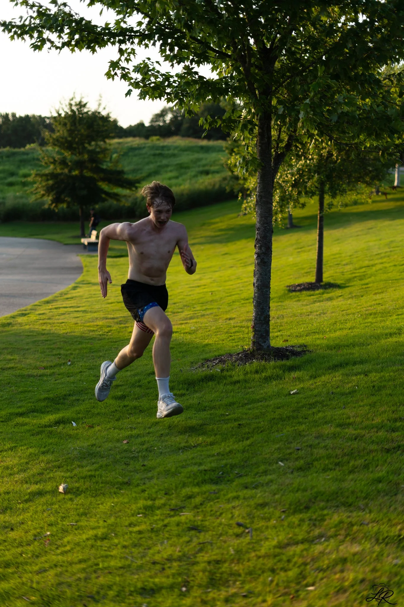 A young man running outdoors in a park with green grass and trees, during sunset. He is shirtless, wearing shorts, white sneakers, and has a joyful expression.