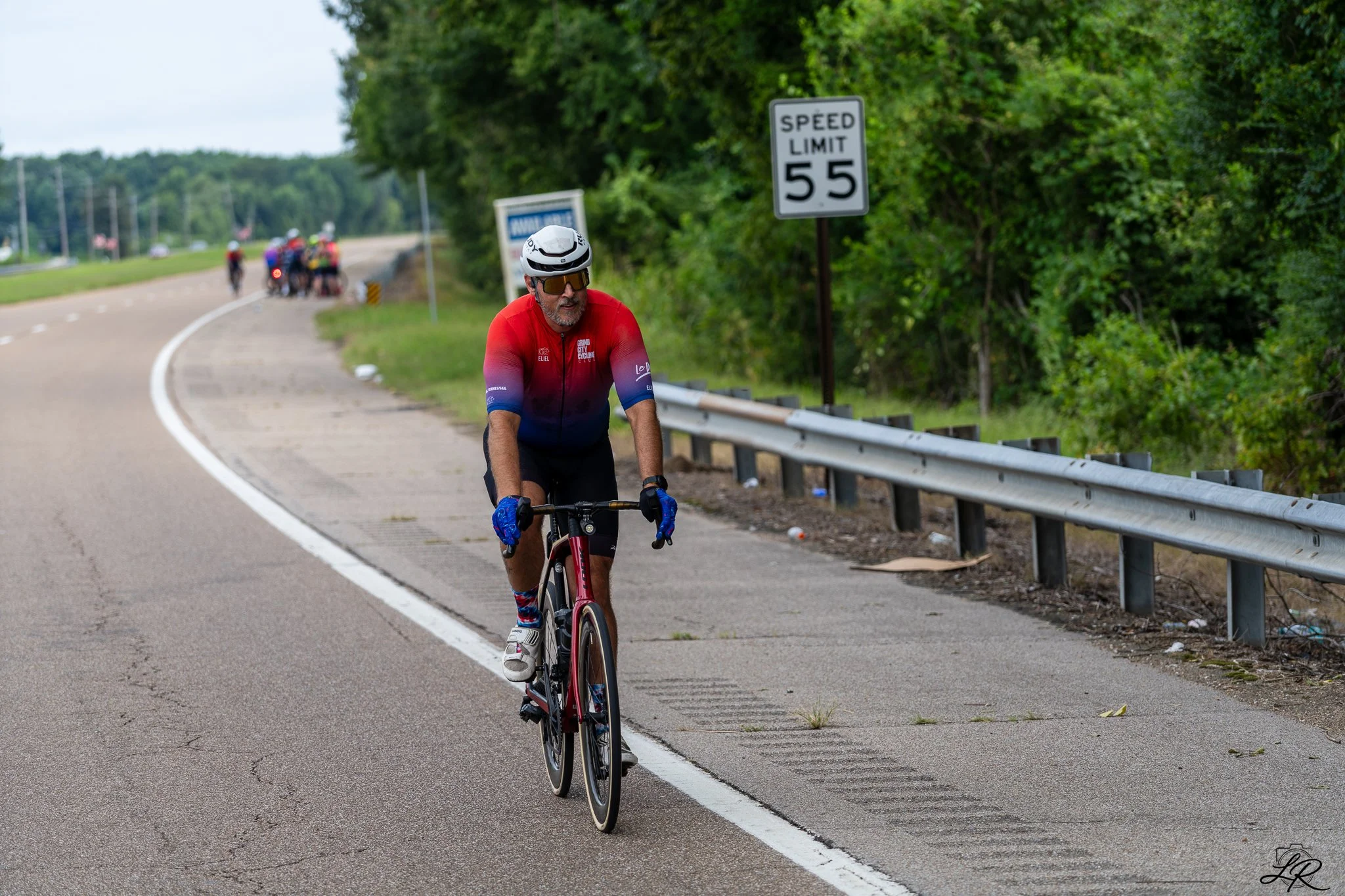 A man rides a bicycle along a road by a guardrail, wearing a helmet and sunglasses. In the background, other cyclists are grouped together. There is a speed limit sign indicating 55 miles per hour and trees lining the road.