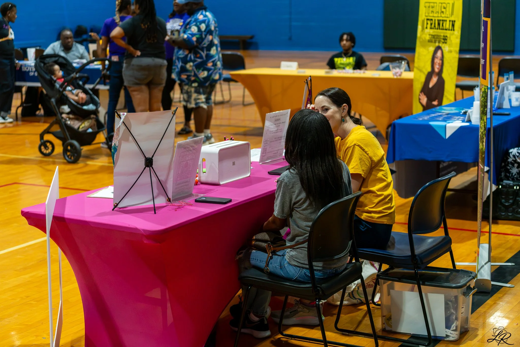 People gathering at a community event or fair inside a gymnasium. There is a pink table with informational materials, and women and children are seen talking and engaging at the booths. A stoller with a child is visible, with banners and tables in th