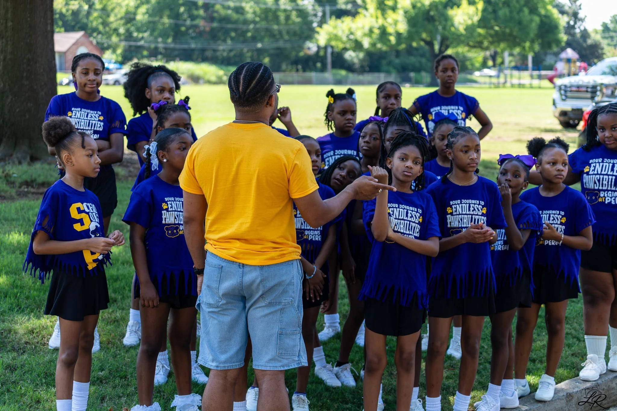 A group of young girls wearing matching blue T-shirts and white sneakers listening attentively to an adult in a yellow T-shirt outdoors in a park.