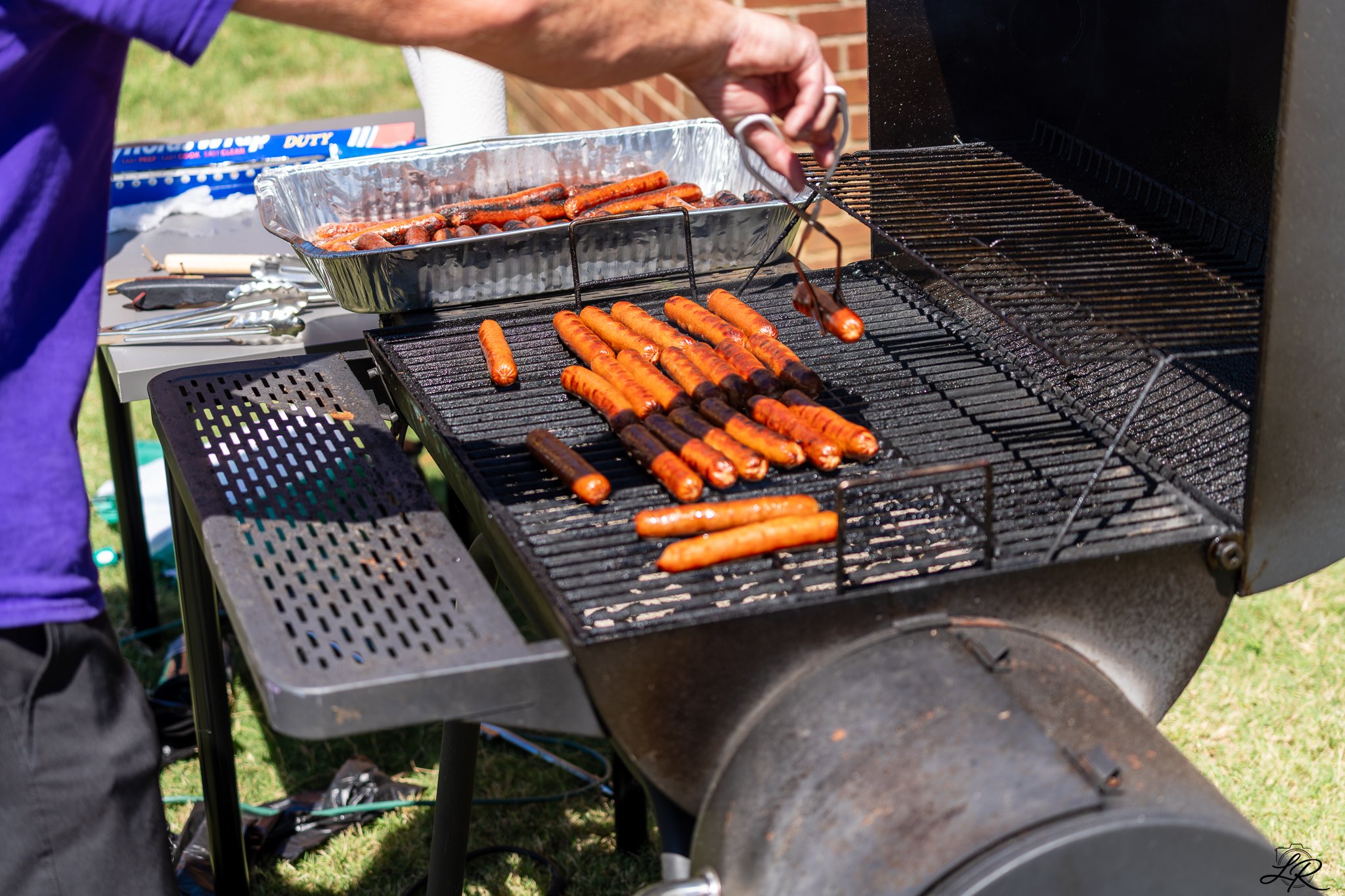 Grill with hot dogs cooking on the grill and in a aluminum tray being rotated by a person.