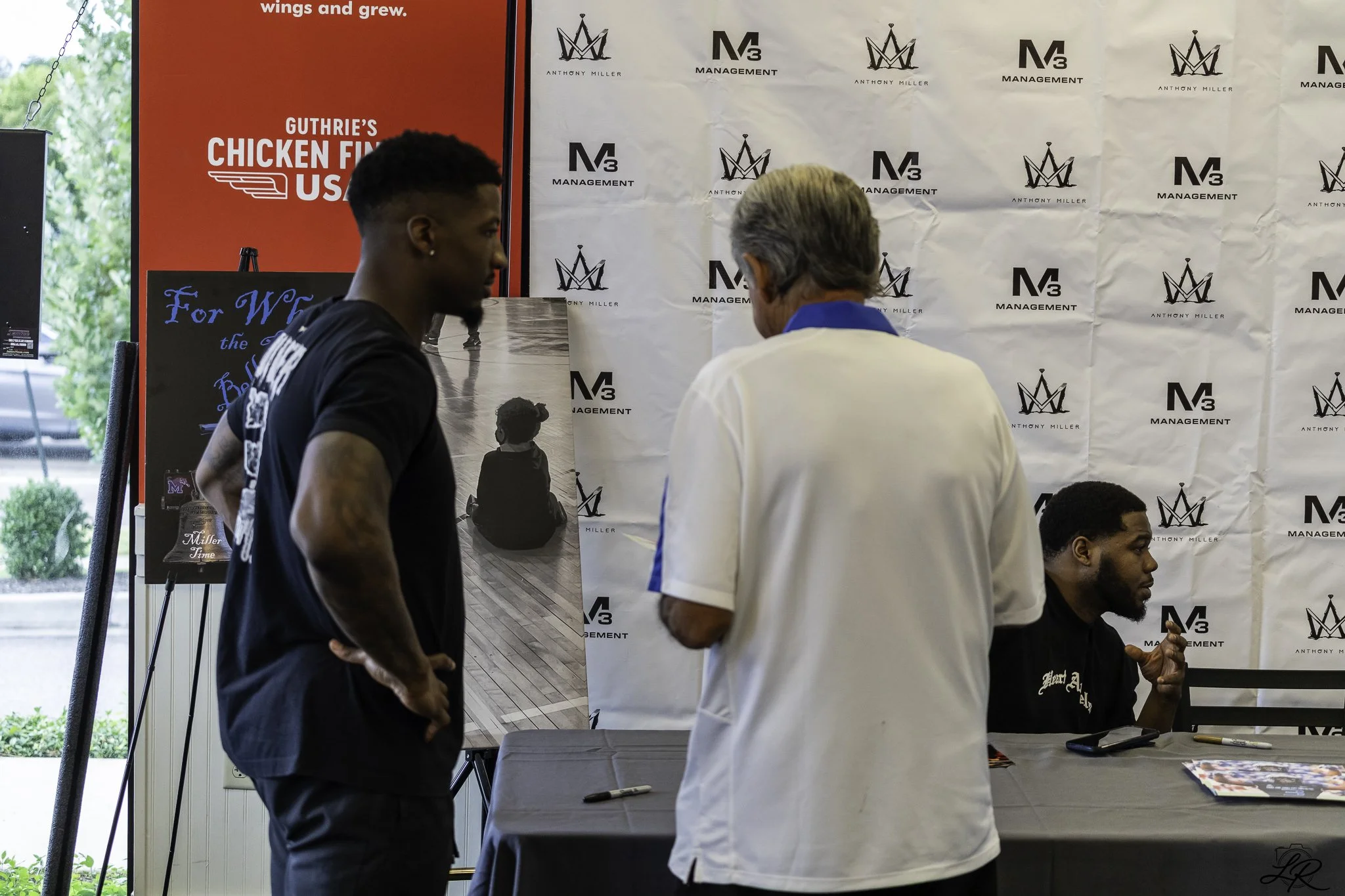 People attending an event with a backdrop featuring logos and promotional posters. A man in a black shirt is in the foreground, and two men are standing near a table where one is seated, engaging in conversation.