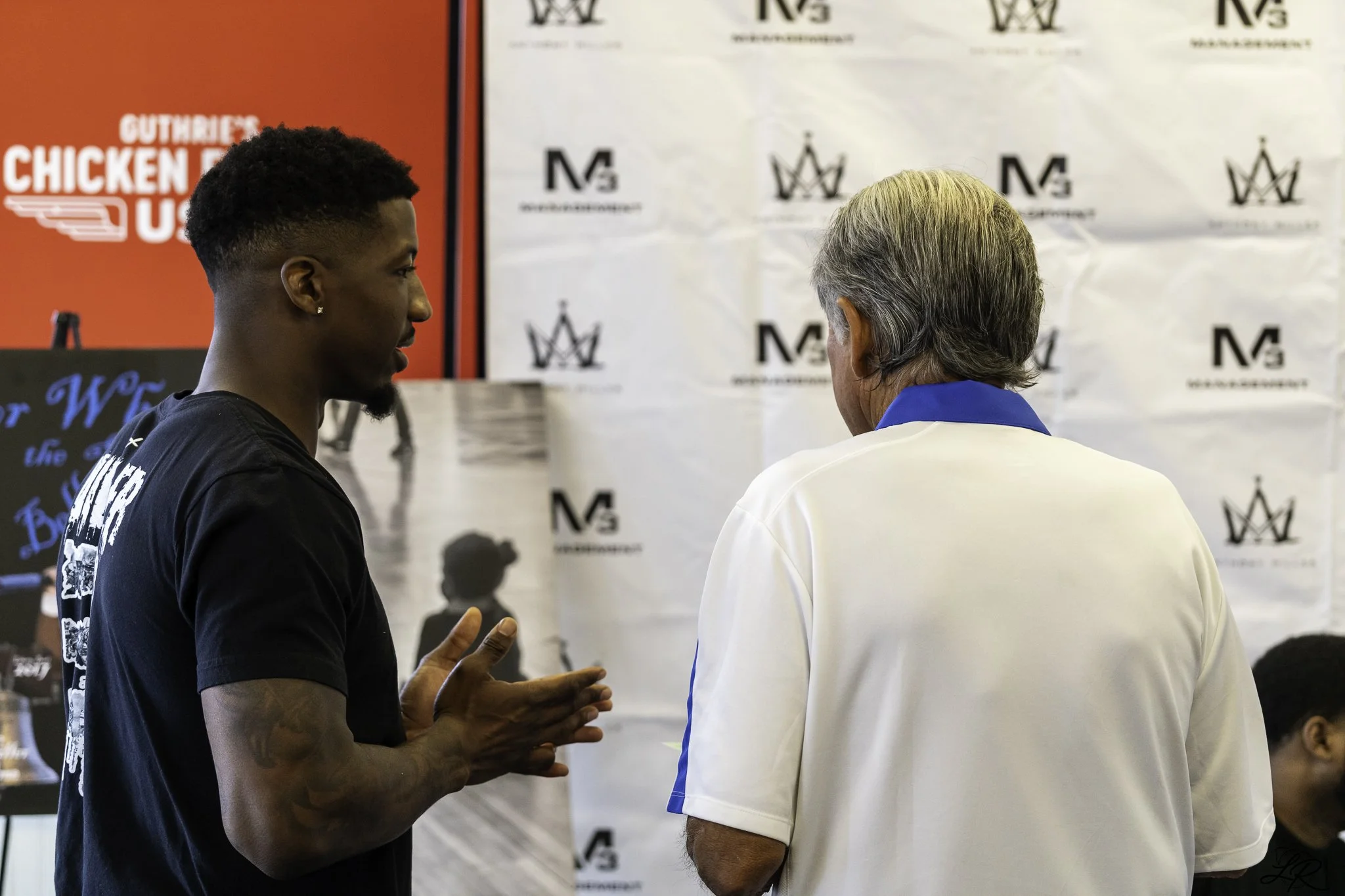 Two men are engaged in a conversation; one has dark skin, short curly hair, a goatee, and wears a black T-shirt with white text, while the other has light skin, gray hair, and wears a white jacket with blue trim. They are standing in front of a white