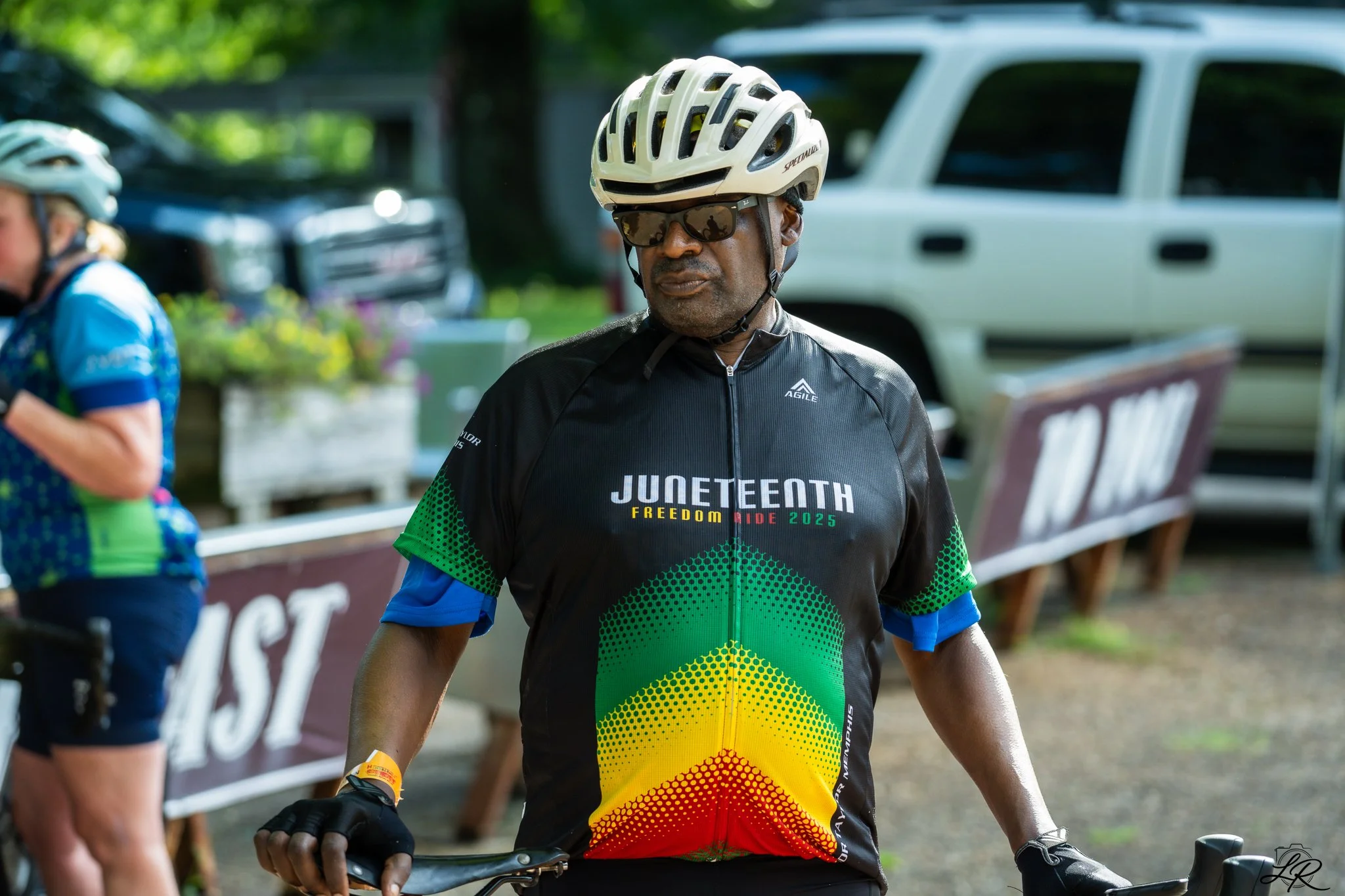 A man wearing a white helmet, sunglasses, and a colorful cycling jersey with rainbow designs stands beside his bike at a cycling event. There are other cyclists and vehicles in the background.